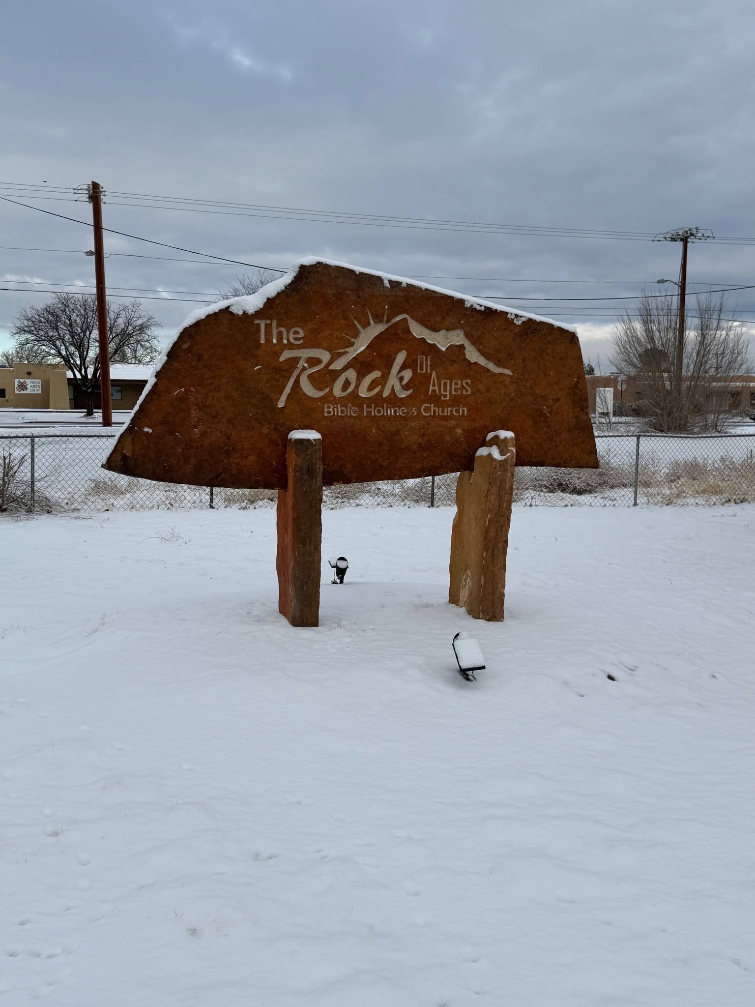 A large wooden sign for 'The Rock of Ages Bible Holiness Church' is displayed in a snowy outdoor setting with a cloudy sky, utility poles, and bare trees in the background.