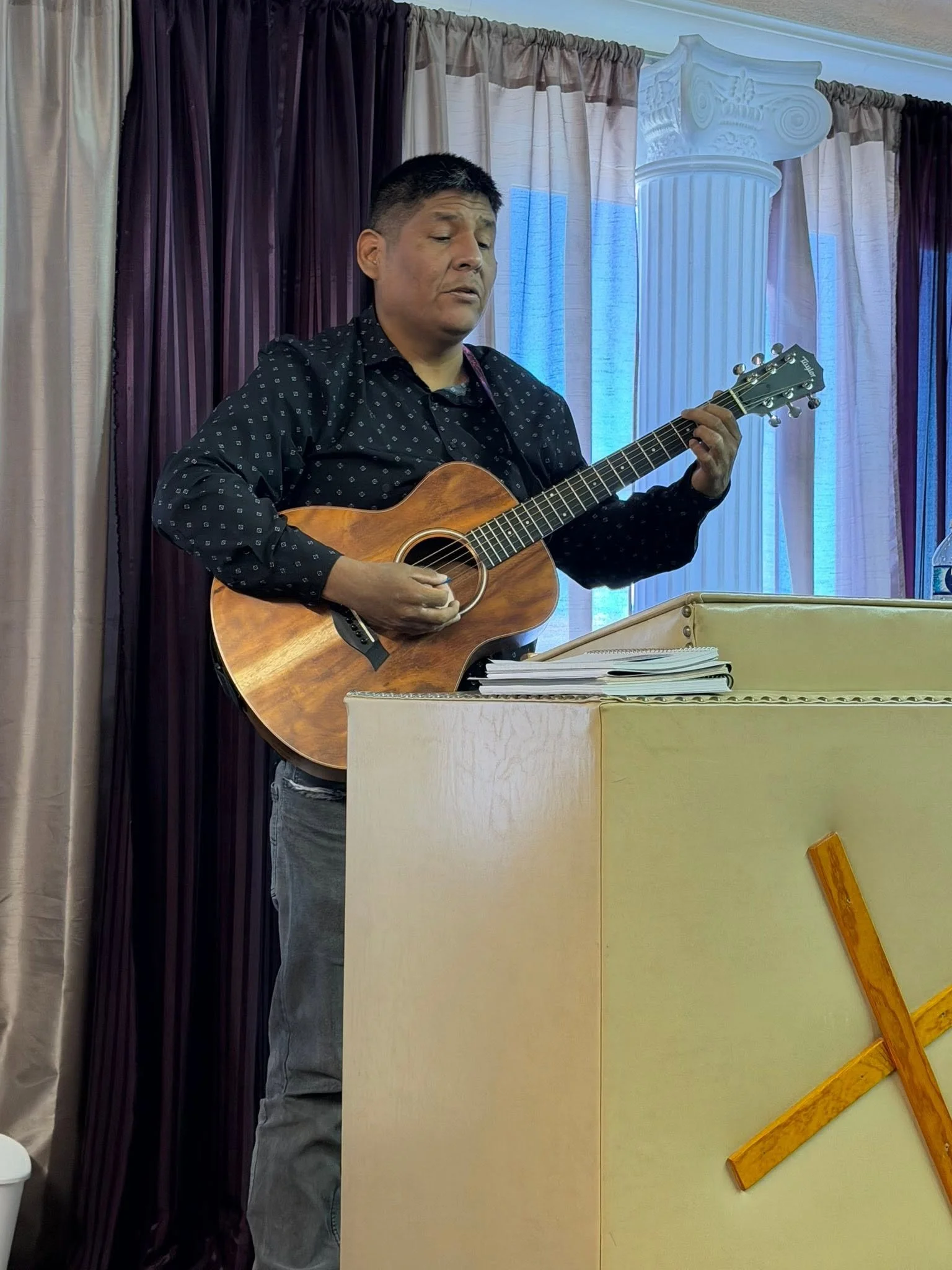 A man playing an acoustic guitar in a room with beige and dark purple curtains, and a white decorative column.