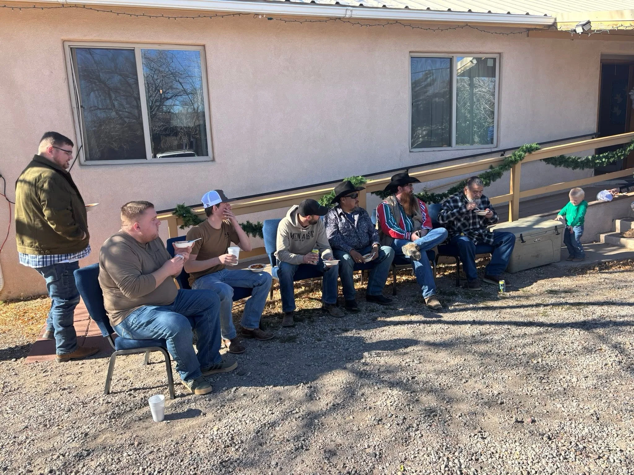 A group of men and a child are sitting and standing on a porch or patio, eating food and drinking from cups. The porch is decorated with Christmas garland and string lights. The background shows a pink stucco house with two windows and a door, and a sunny outdoor environment.