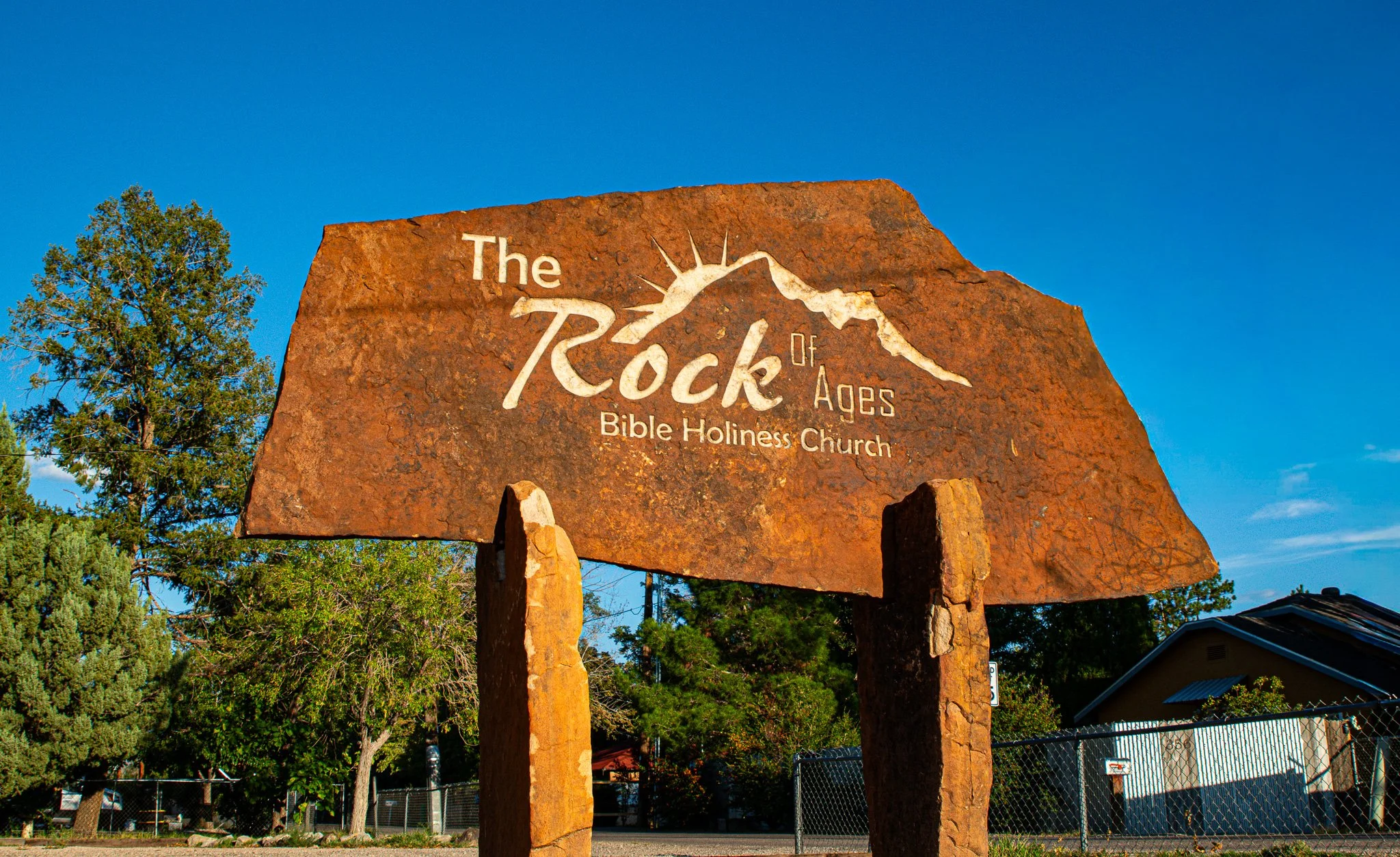 A large engraved stone sign reading 'The Rock of Ages Bible Holiness Church' with a mountain range graphic, set against a clear blue sky and surrounded by trees and a chain-link fence.