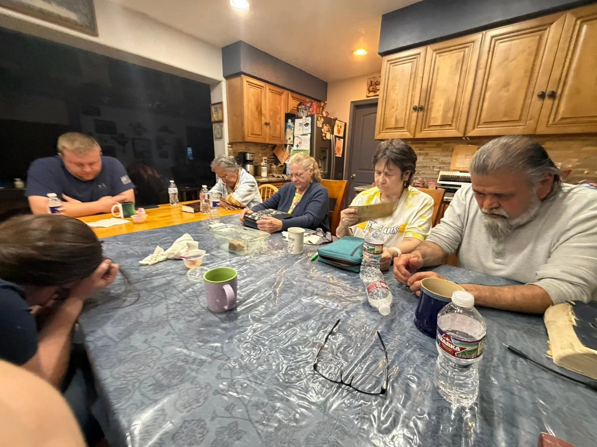 A group of people sitting around a dining table in a kitchen, with various food items, bottles of water, cups, and personal belongings on the table. The kitchen has wooden cabinets and a brick backsplash.