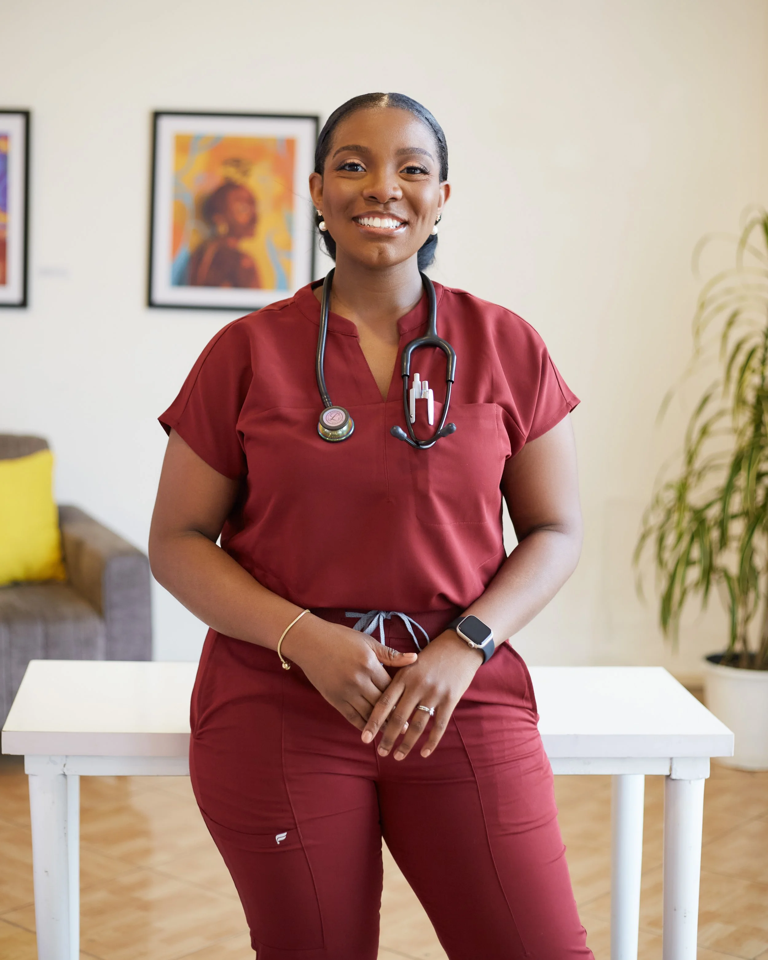 A smiling woman wearing a maroon medical uniform with a stethoscope around her neck, standing indoors in front of a white table and artwork on the wall.