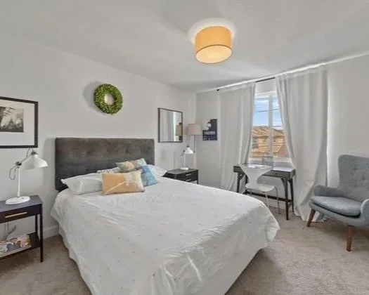 An organized bedroom with a bed, nightstands, and lamps, a dresser, a window with white curtains, a desk with a chair, and an armchair.