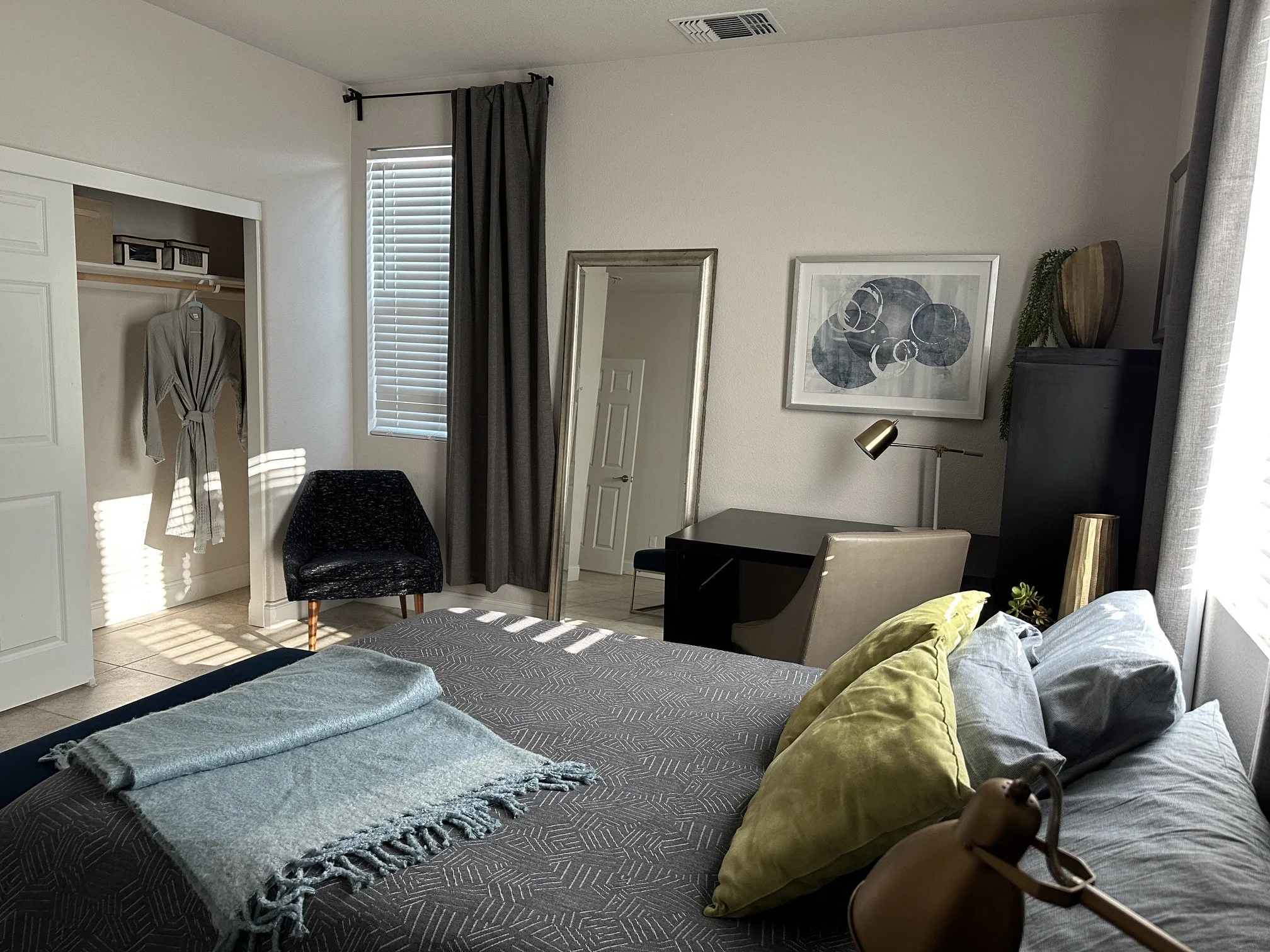 Organized bedroom with bed, pillows, and blanket in foreground, desk with chair, artwork on wall, mirror, clutter-free closet with robe, window with blinds and curtains, black chair, and decorative items on a cabinet.
