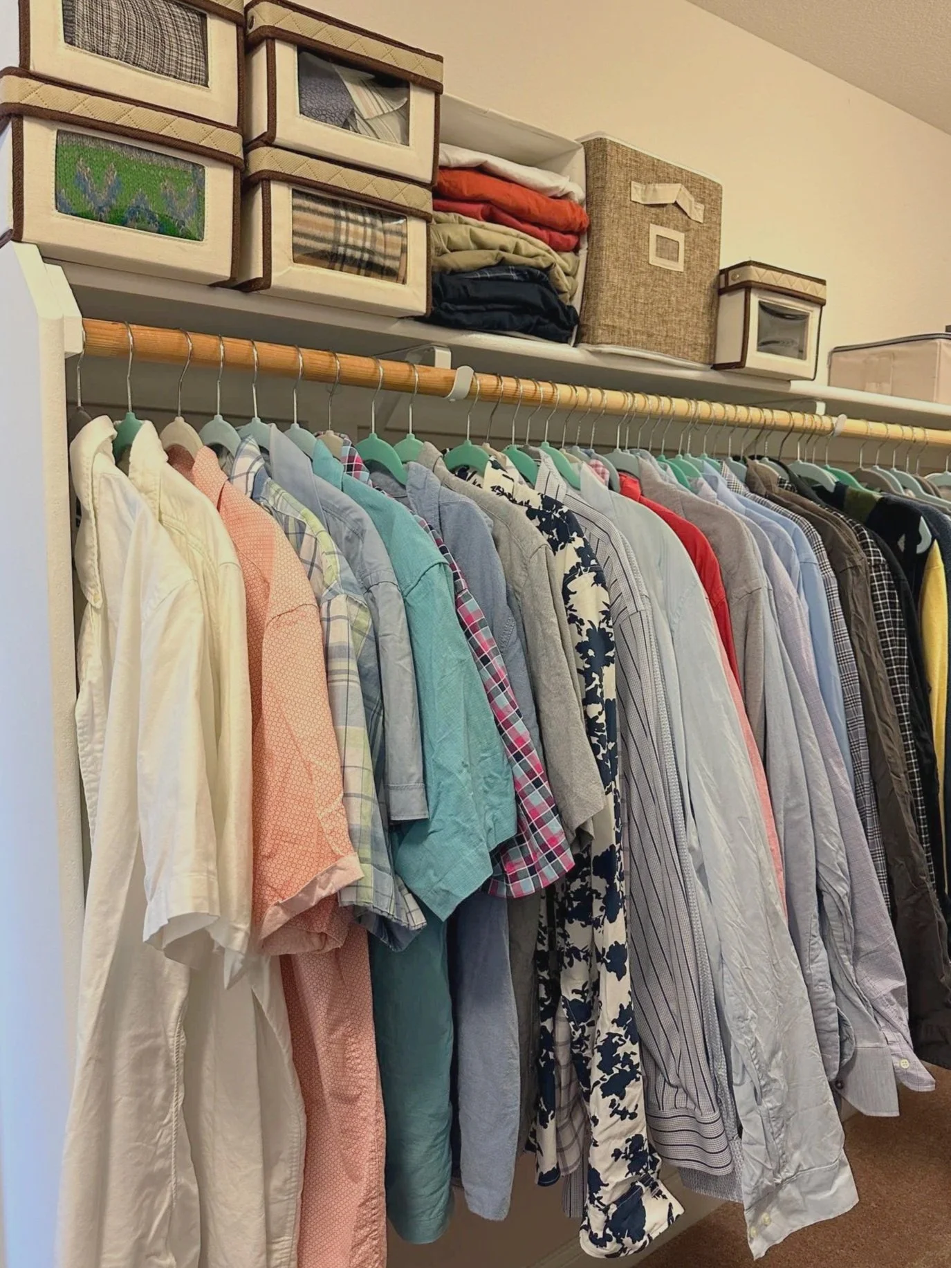 A clutter-free closet with various men's shirts organized in different colors and patterns, with storage boxes and folded clothes on a shelf above.