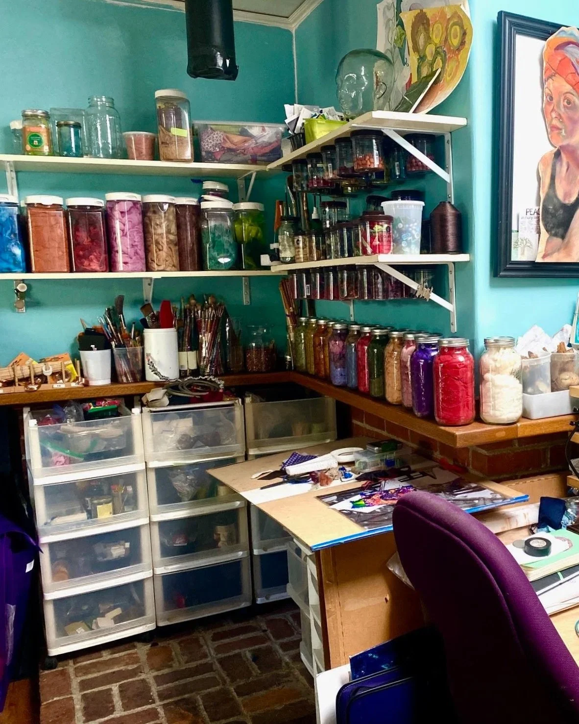 An organized artist's workspace with shelves of jars containing colored materials, art supplies such as brushes and paints on a desk, and artwork hanging on turquoise walls.