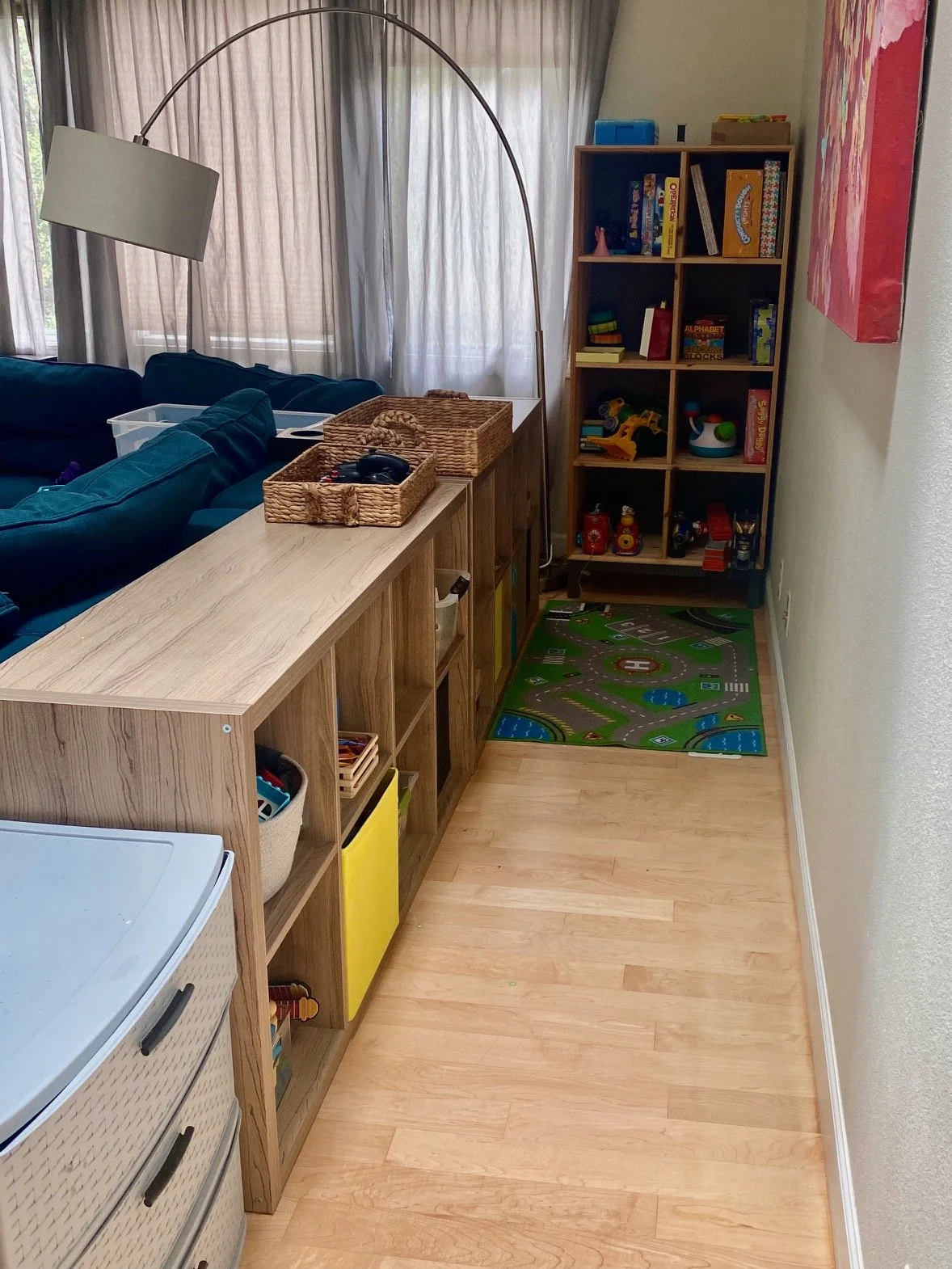 Living room or playroom area with an organized wooden shelf holding toys and books, a colorful playmat on the floor, and a large window with curtains in the background.