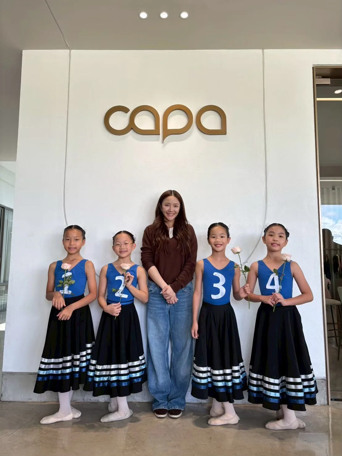 Group of five young girls and one woman standing in front of a white wall with the word 'capa' on it; the girls are dressed in matching blue and black outfits with white stripes, each holding a pink rose, and numbered 1 through 4, standing in a line.