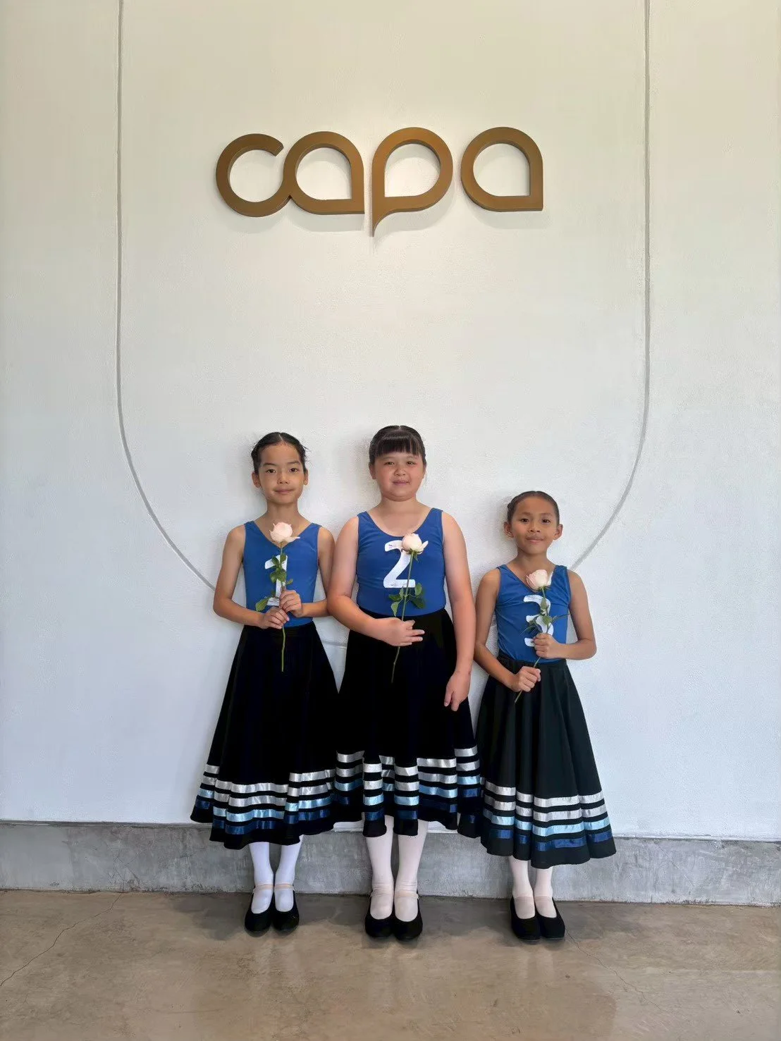 Three young girls in black skirts and blue tops holding pink roses standing in front of a white wall with the word 'capa' displayed above them.