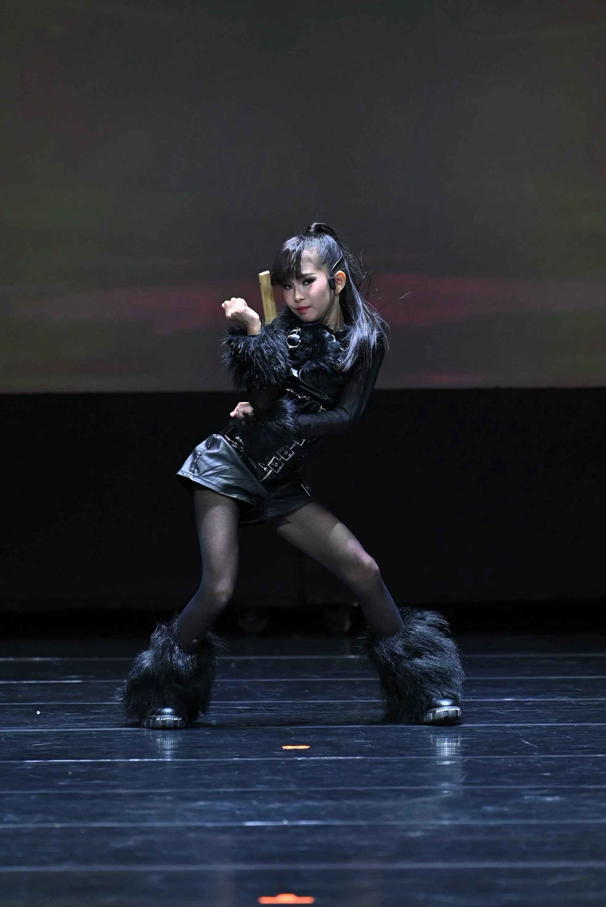 A woman on stage dressed in black clothing, furry accessories, and boots with fur, striking a pose with one arm bent and the other holding a prop, against a dark background.