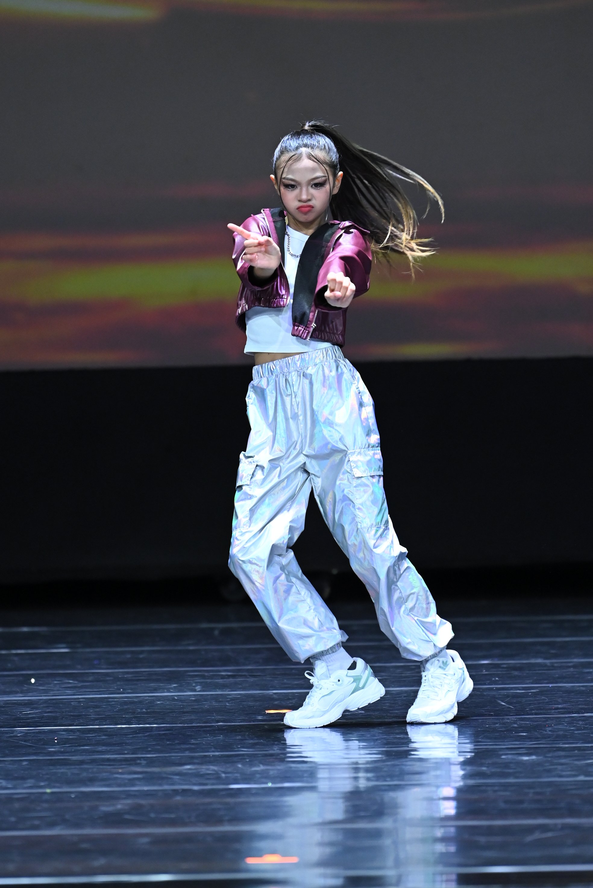 Young girl dancing on stage, wearing shiny white pants, a white shirt, a purple jacket, and white sneakers, with long hair in a high ponytail.