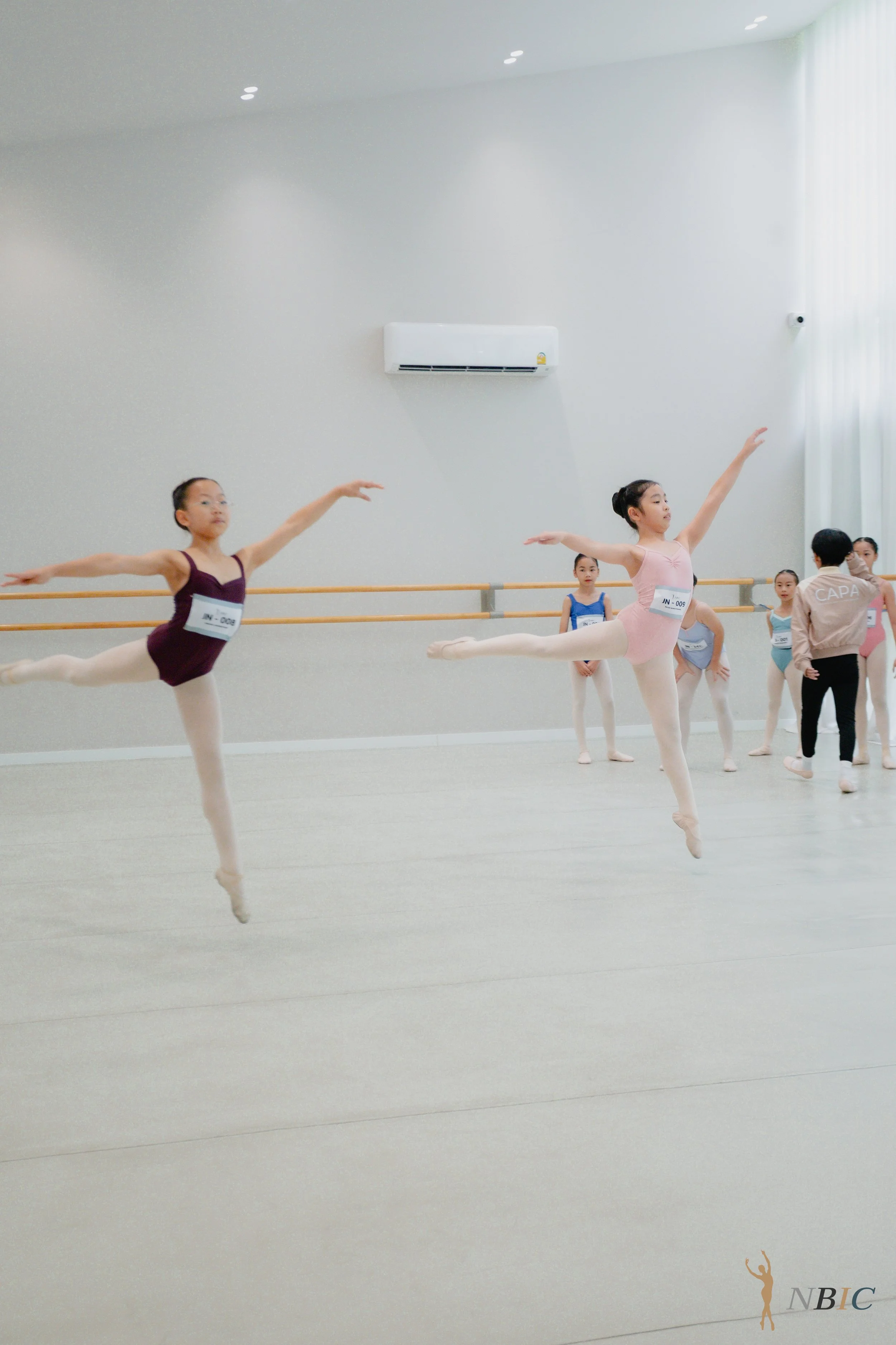 Young female ballet dancers practicing in a dance studio with white walls, wooden ballet barres, and a large window with curtains. The dancers are wearing leotards and tights, performing a pose with one leg extended behind and arms outstretched.