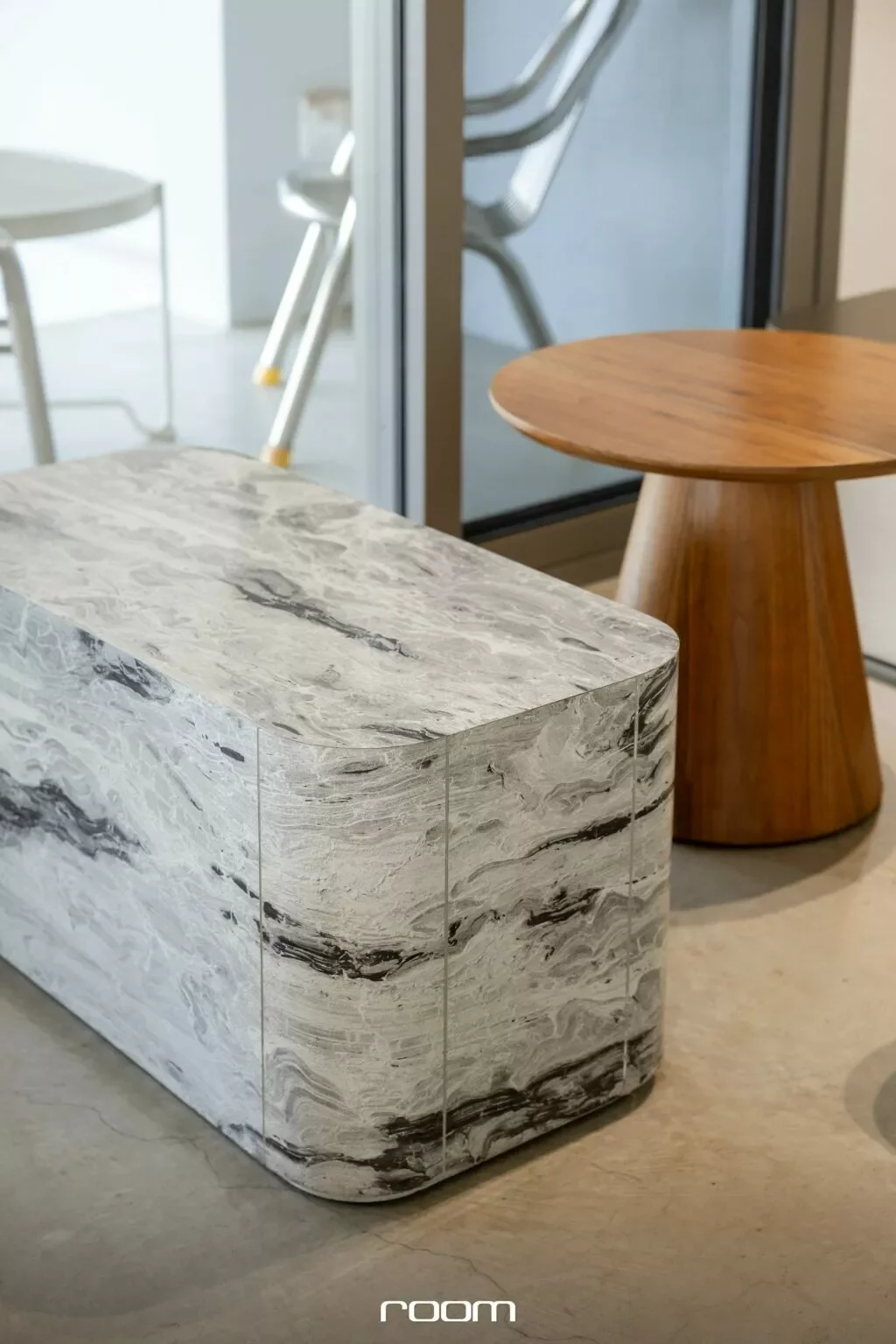 Close-up of a marble coffee table with swirling gray and white patterns, next to a wooden side table. In the background, there are chairs and a glass door leading to another room.