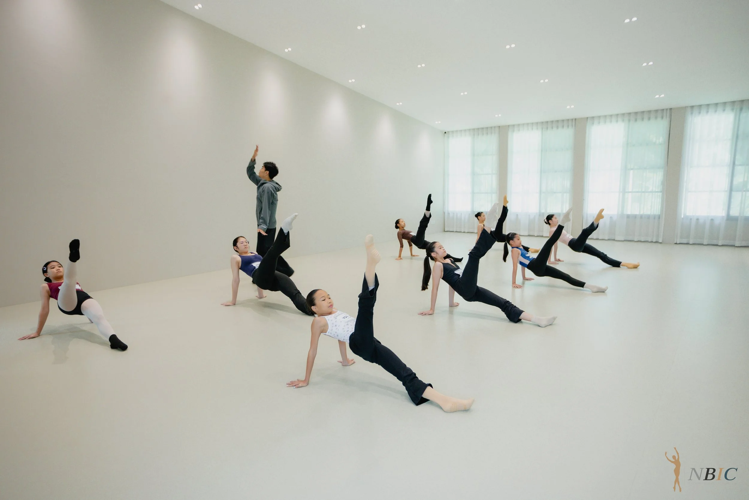 A group of young ballet dancers practicing their routine in a dance studio with large windows, wearing ballet attire, while an instructor guides them in poses and stretches.