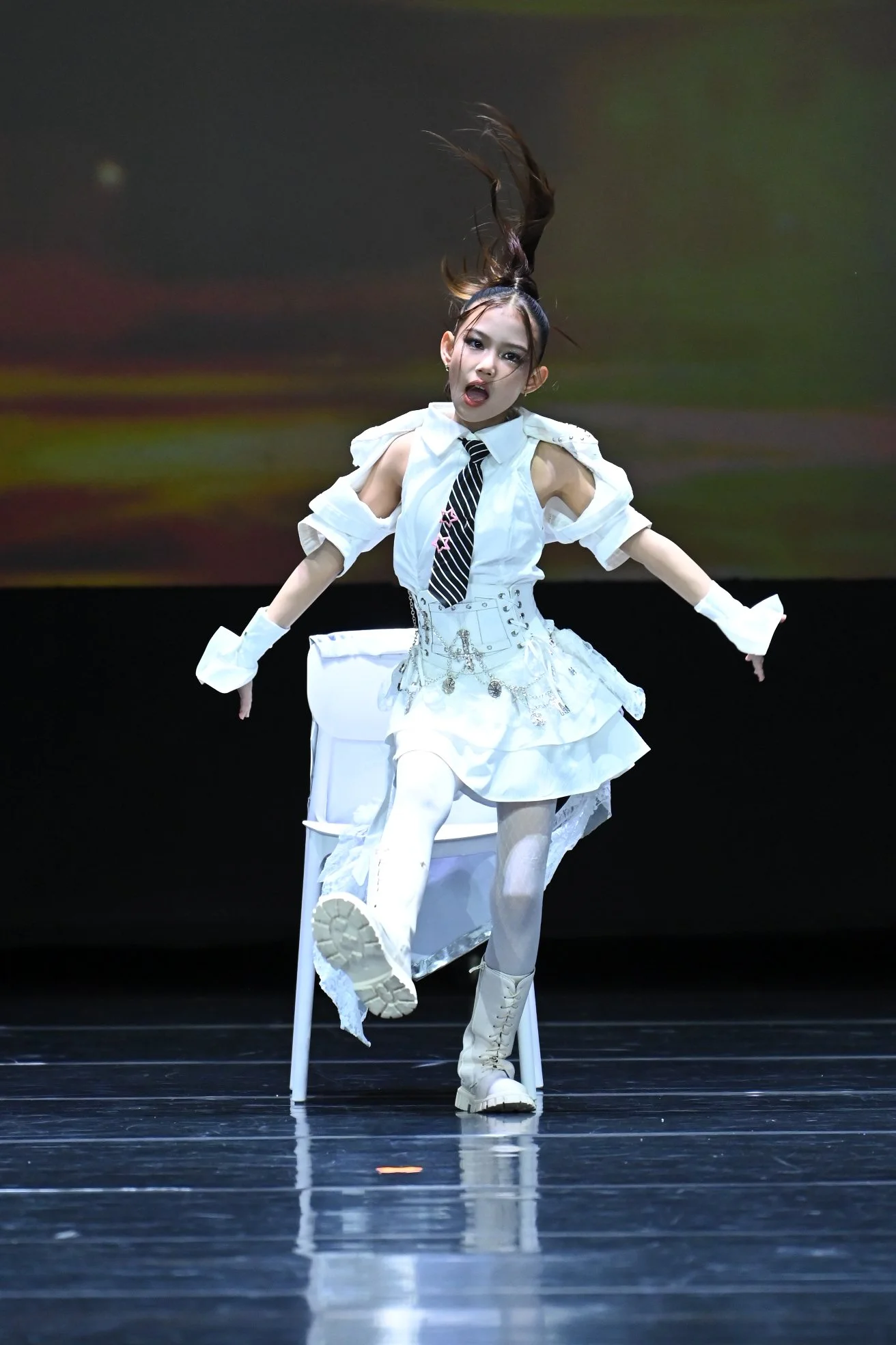 A performer dressed in a white outfit with a schoolgirl style, black and white striped tie, white gloves, and knee-high boots, is sitting on a transparent chair on a stage. She has long hair styled in a high ponytail and appears to be in a dance or t