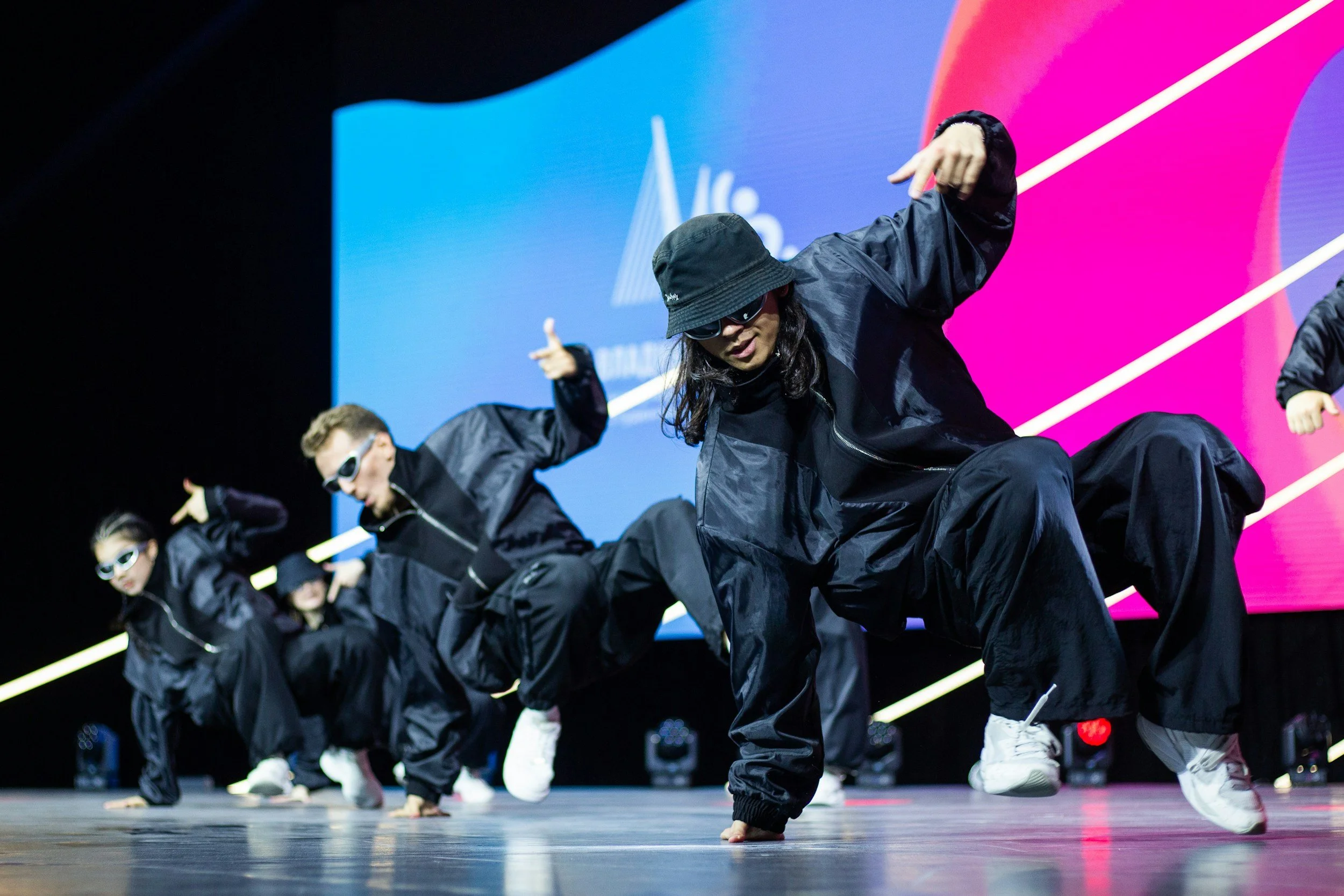 A group of dancers on stage performing a breakdance routine, dressed in black sportswear and sunglasses, with a colorful background and stage lights.