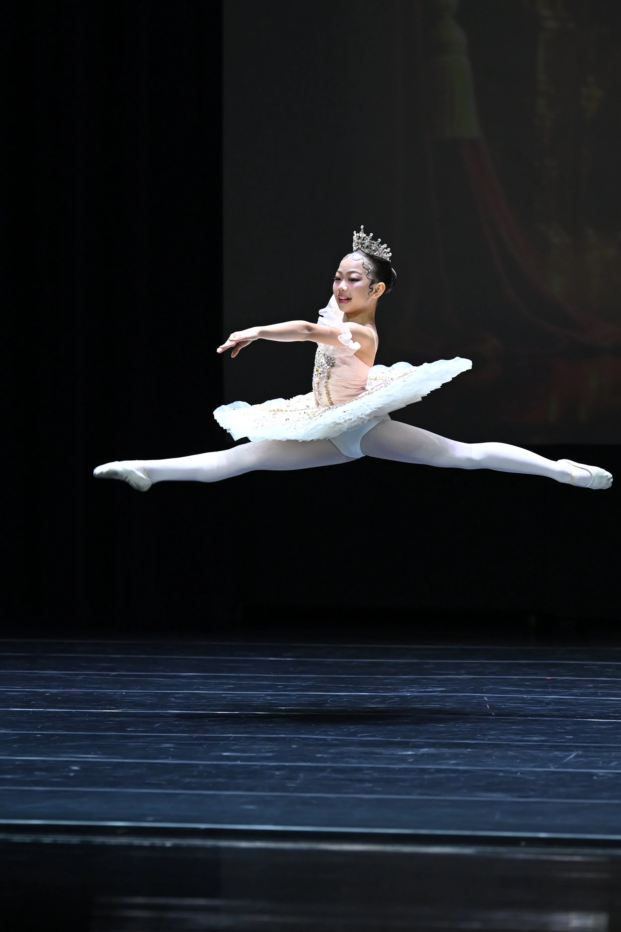 A young ballet dancer performing a grand jeté on stage, dressed in a white tutu with a tiara, with a dark background.