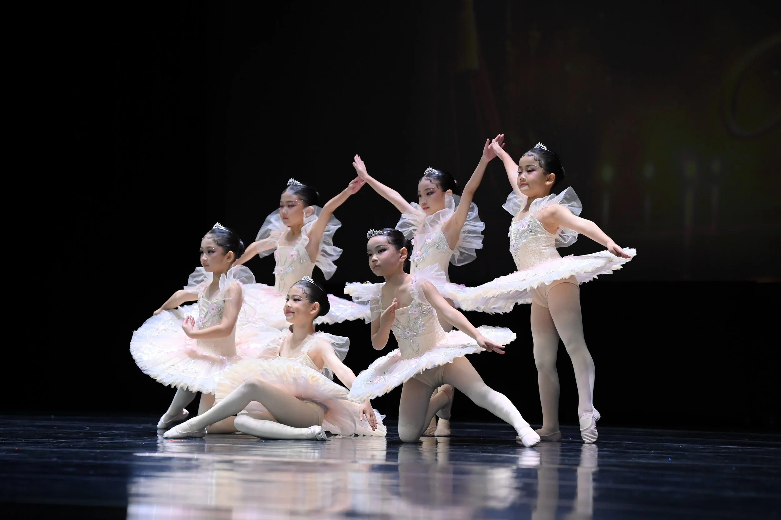 A group of young ballet dancers in white tutus and tiaras performing on stage, with some dancers kneeling and others standing with arms raised.