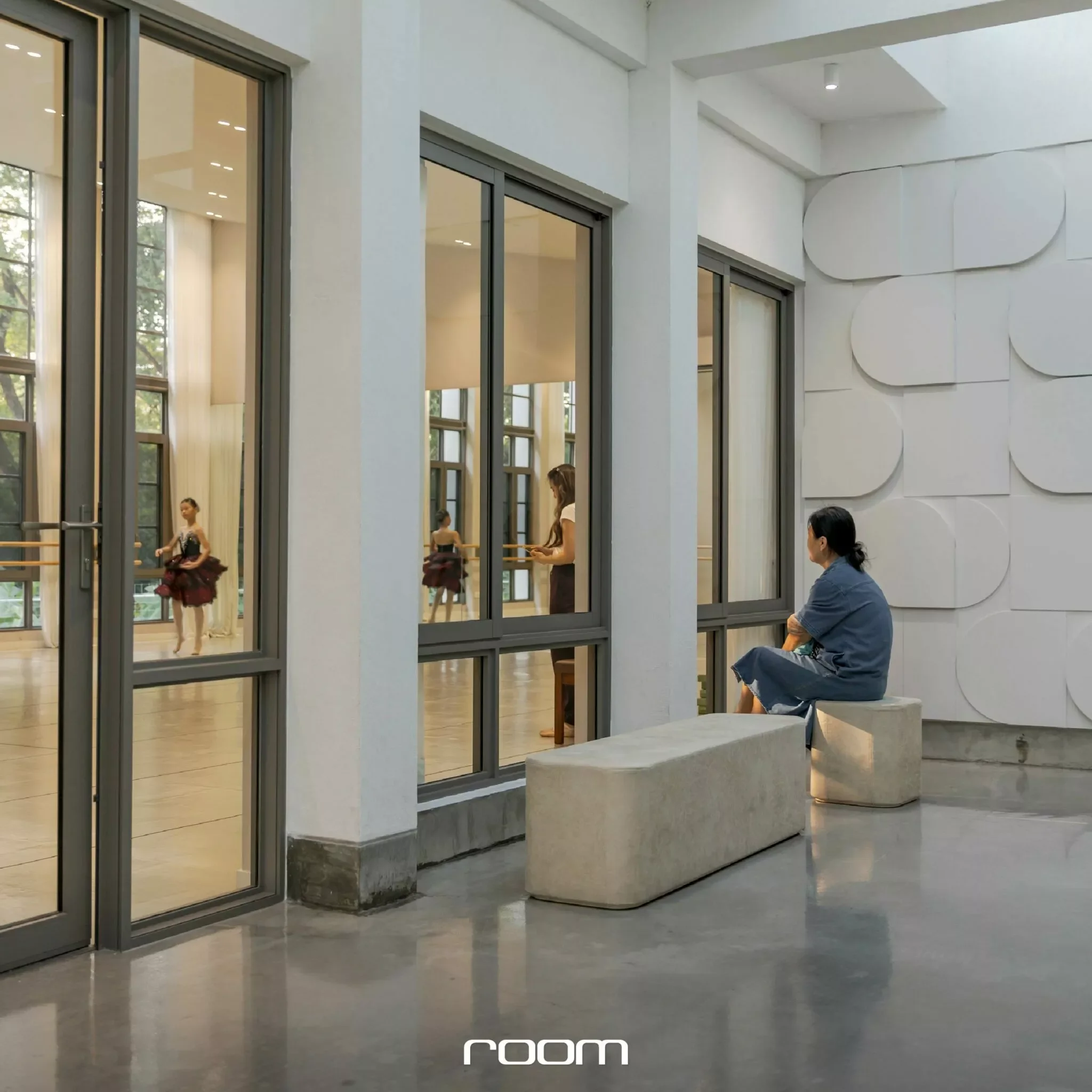 A modern indoor space with large floor-to-ceiling windows and white walls. Two young ballet dancers are practicing in front of a mirror, dressed in ballet attire. An older man is seated on a bench near the wall, observing or resting.