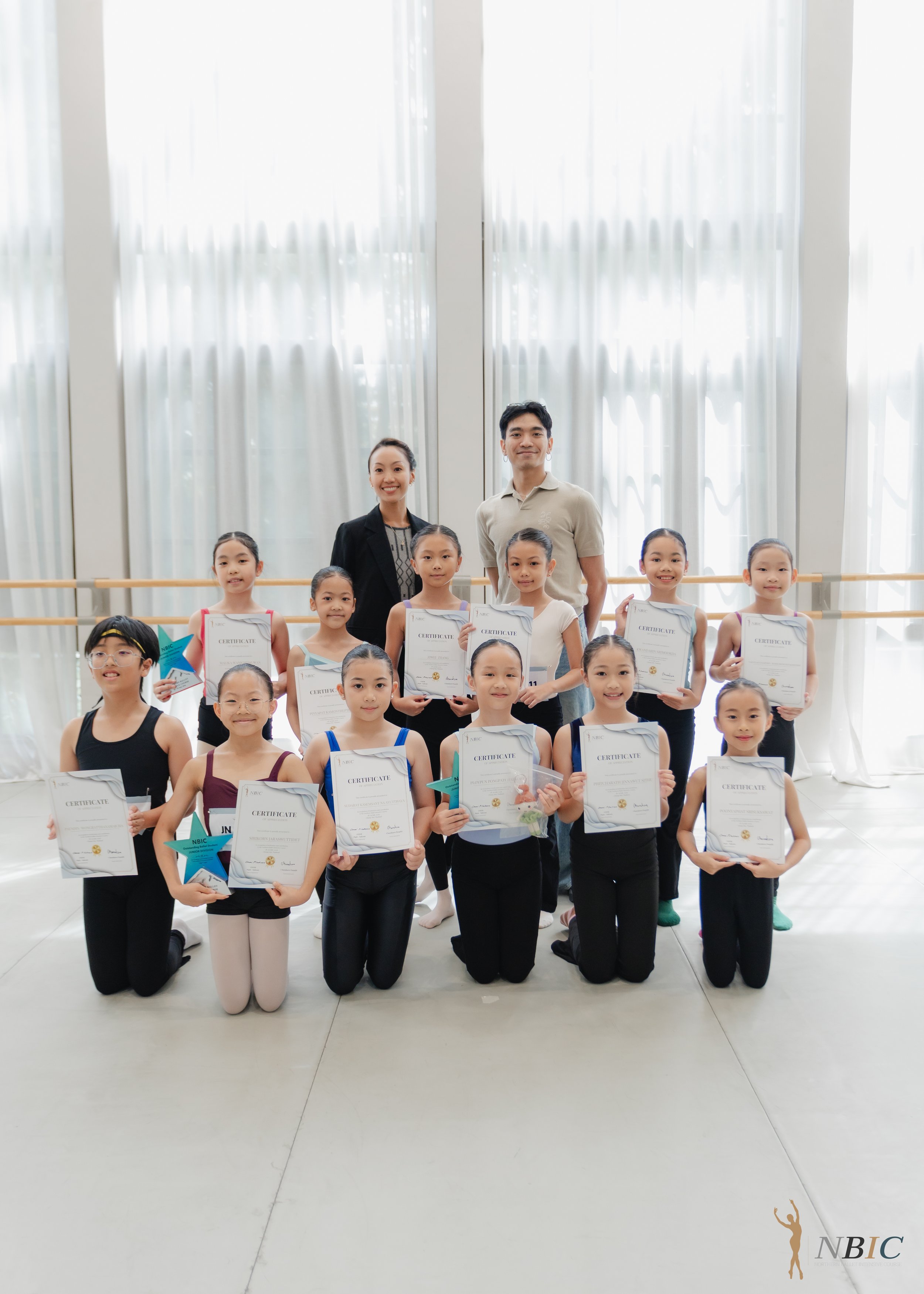 Group of young girls and two adult instructors in a dance studio, holding certificates of achievement. The students are dressed in dance attire and are smiling. Large windows with sheer curtains are in the background, and a ballet barre runs across t