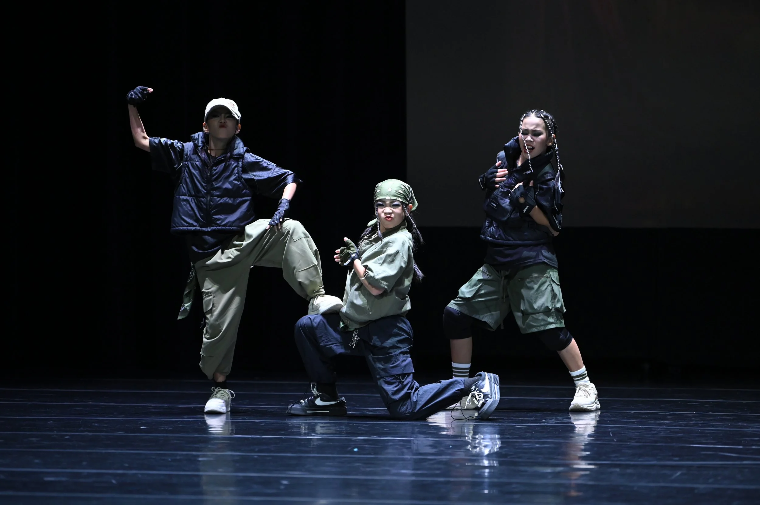 Three dancers perform on stage against a dark background. The dancers are dressed in streetwear, including oversized pants, jackets, gloves, and accessories, striking dynamic poses with expressive facial expressions.