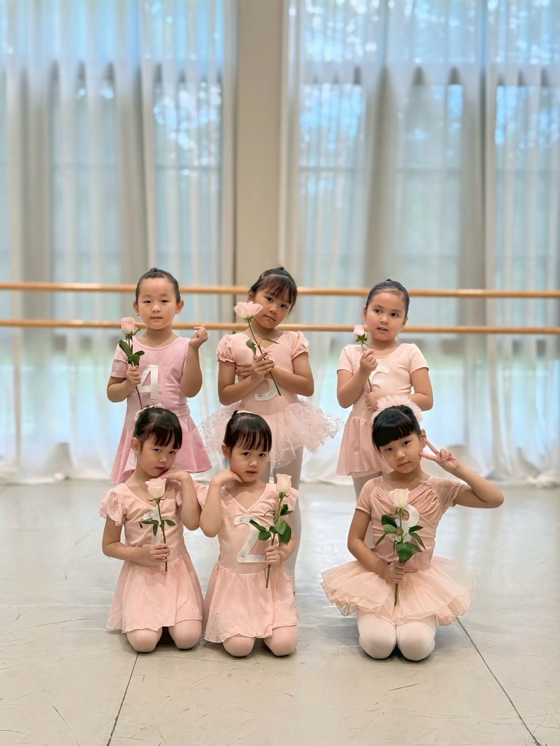 Six young girls dressed in pink ballet costumes, holding roses, standing in a dance studio with a barre and large windows with curtains in the background.