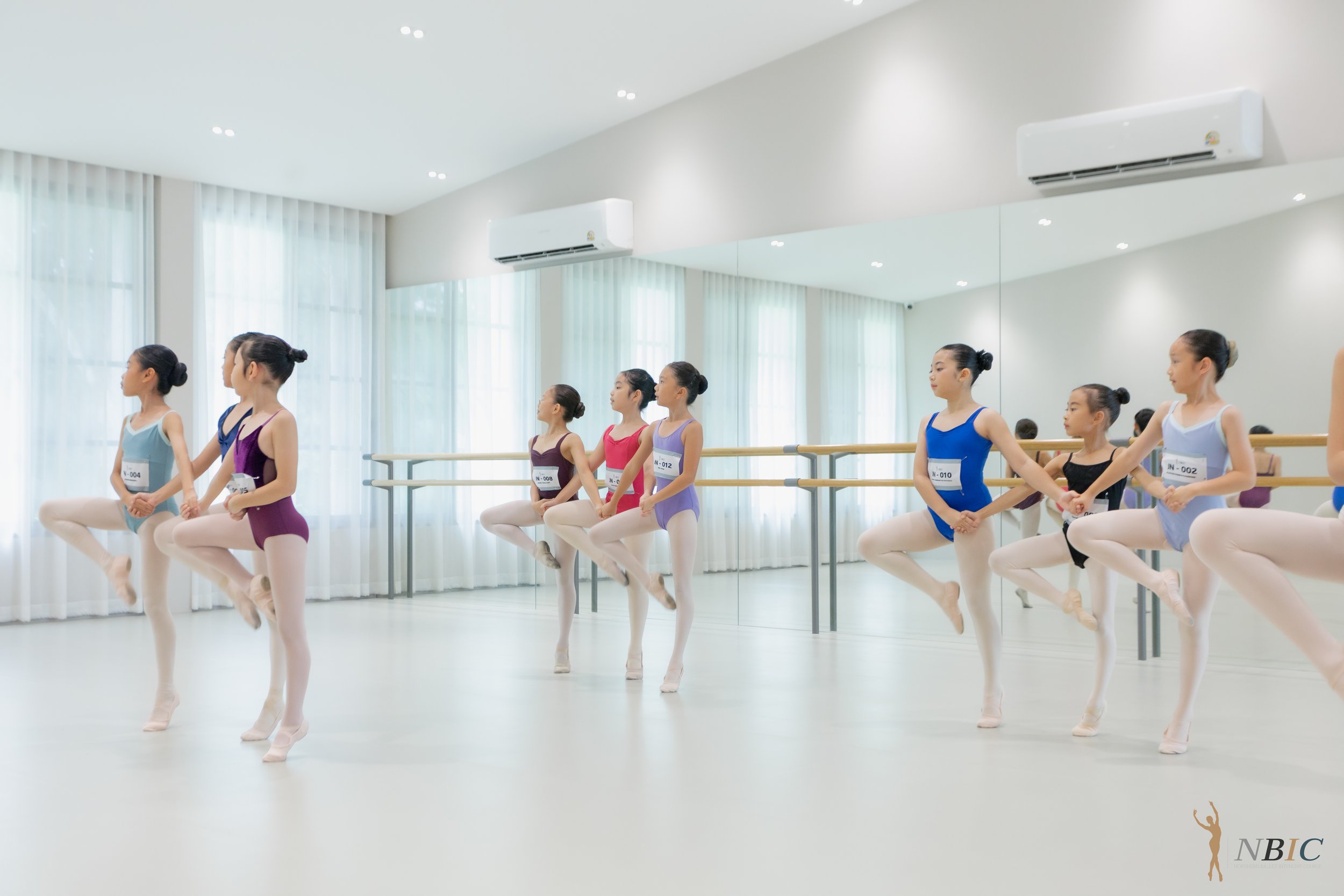 Young ballet dancers in colorful leotards and tights practicing at a ballet barre in a bright dance studio with mirrored wall and large windows.