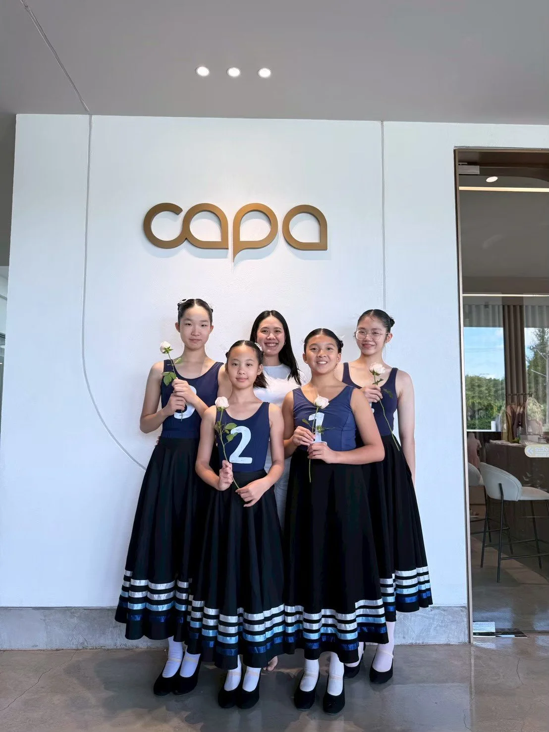 Group of young girls in black and navy blue dance costumes holding white roses standing with their instructor in front of a white wall with a gold logo reading 'capa' at a dance studio.