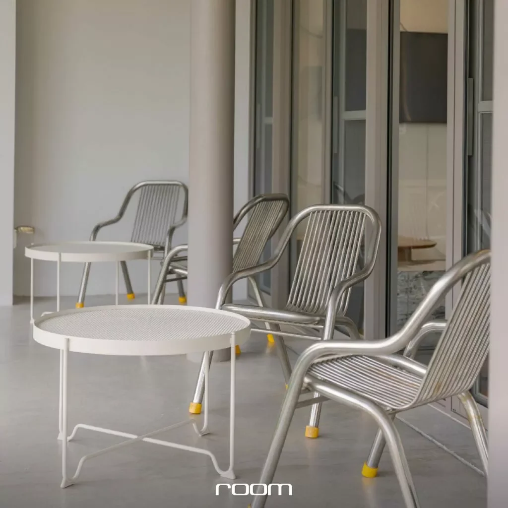 Four metal patio chairs with armrests and yellow tips on their legs are positioned next to a white, round metal table with a perforated surface on a gray concrete floor in front of a building with glass sliding doors.