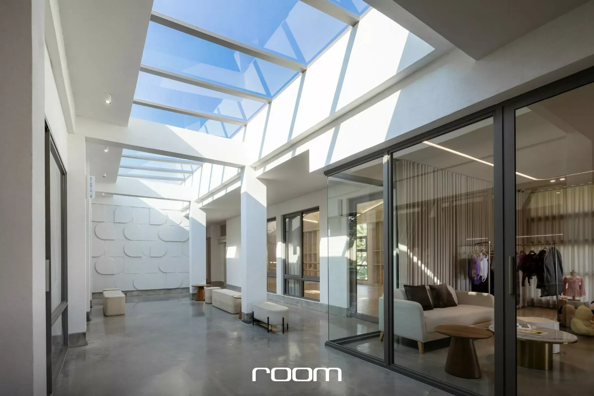Modern interior with a glass ceiling, white walls, and large sliding glass doors opening into a room with clothing displays and a sitting area.