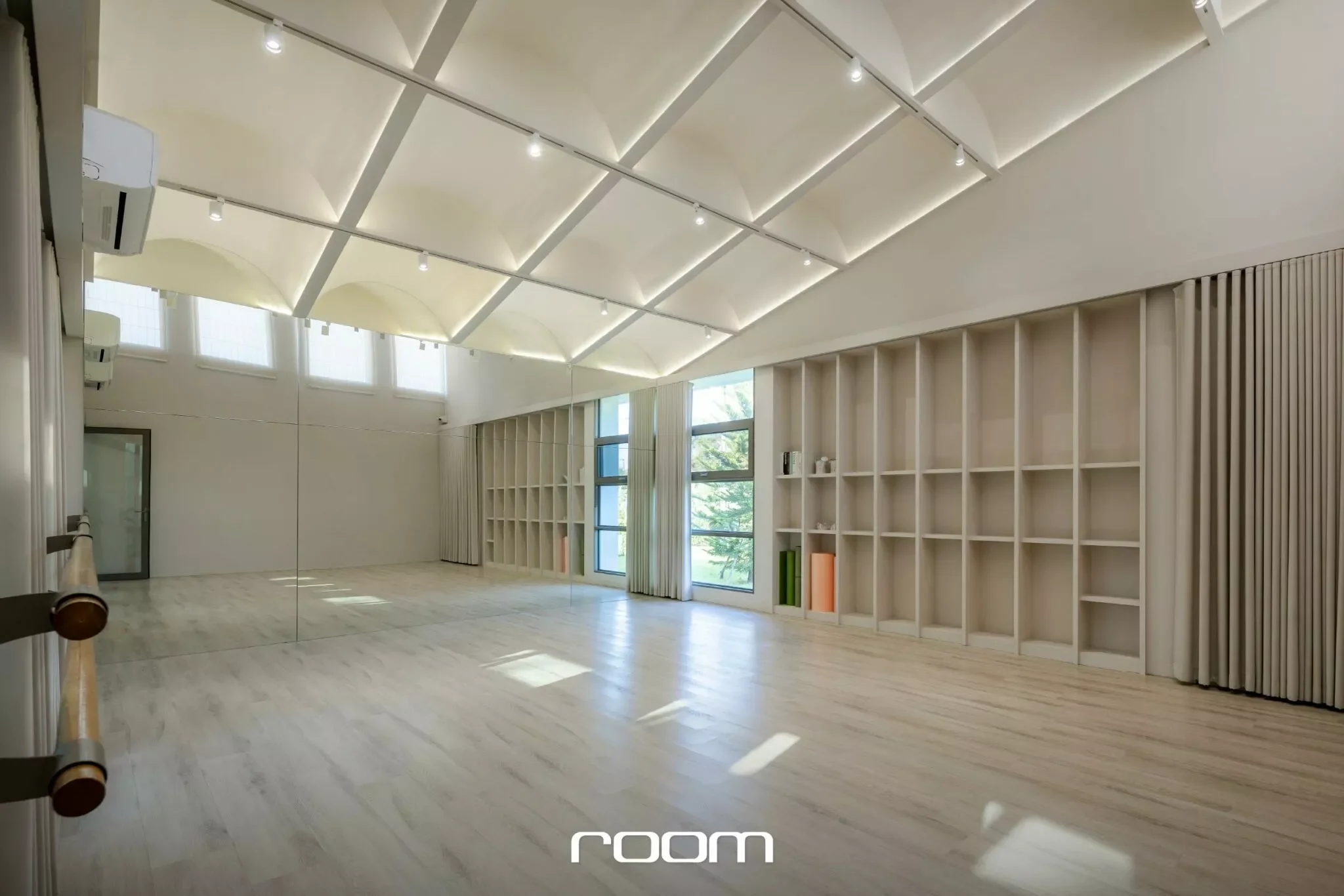 A spacious, well-lit room with a high, vaulted ceiling featuring white panels and small spotlights. Large windows with sheer curtains allow natural light to fill the space. The floor appears to be light-colored wood or laminate, and there is a wall w