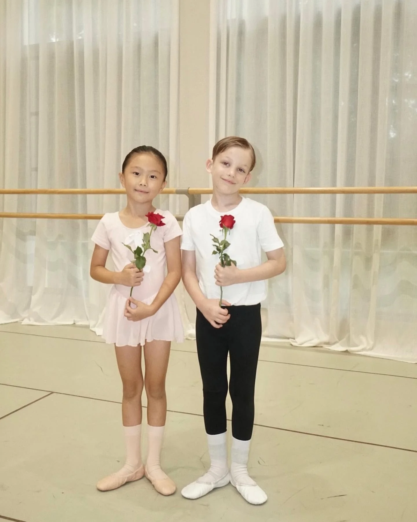 Two young ballet dancers, a girl and a boy, standing side by side in a dance studio, each holding a red rose and smiling at the camera. The girl is wearing a light pink ballet dress, tights, and slippers, and the boy is wearing a white t-shirt, black pants, and ballet slippers. The background features a ballet barre and sheer curtains.