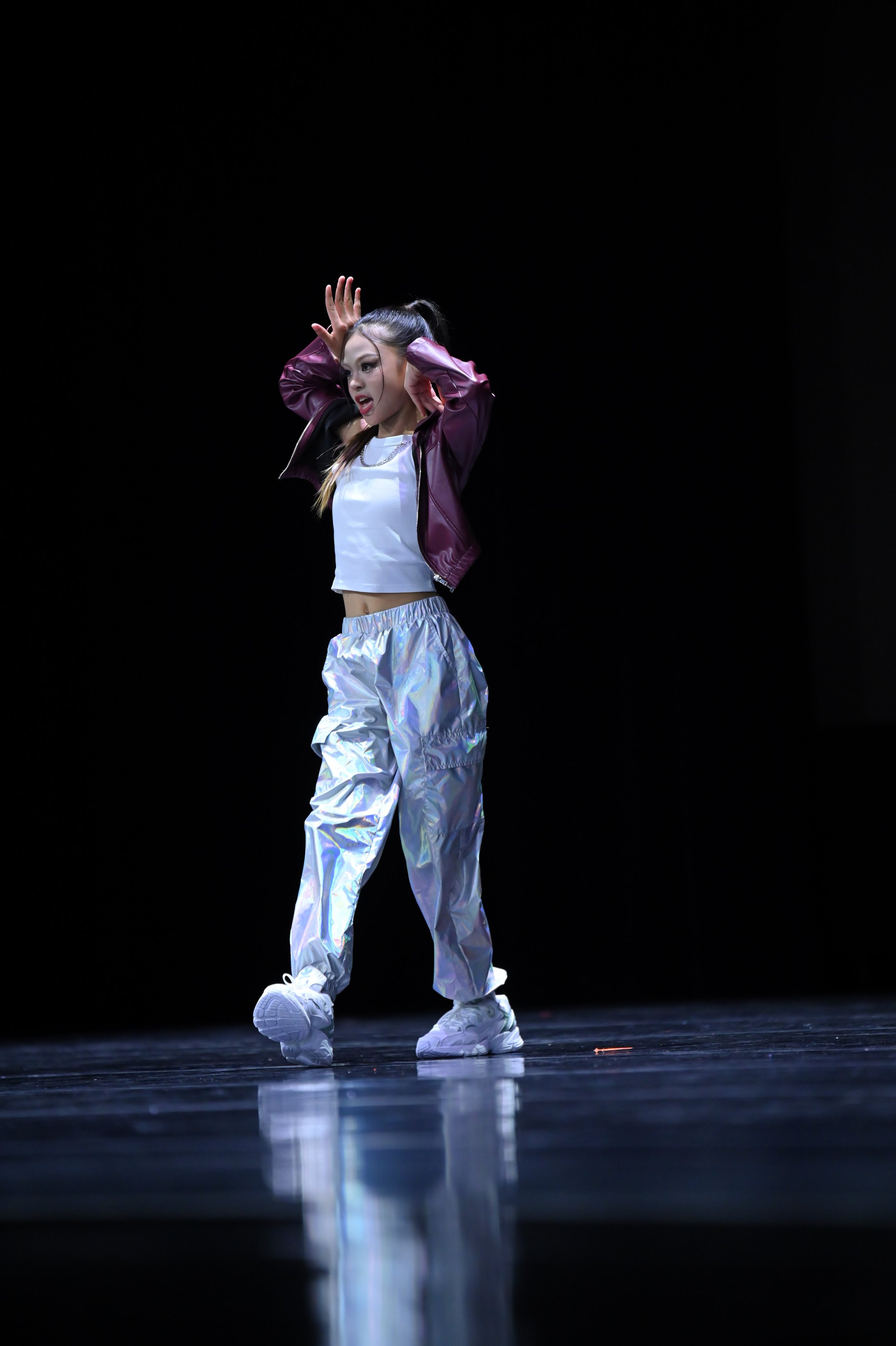 A young female dancer performing on stage, dressed in a shiny purple jacket, white crop top, iridescent pants, and white sneakers, with a dark background and a reflection on the floor.