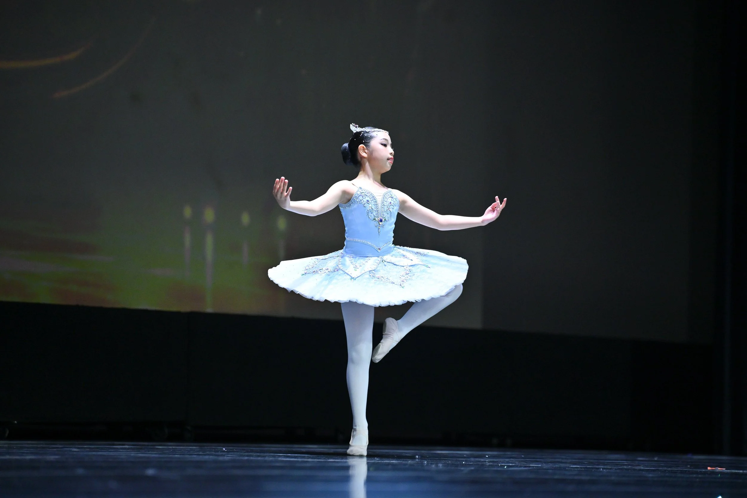 A young ballet dancer practicing on stage, balancing on one leg with arms outstretched and wearing a blue tutu with intricate embellishments.