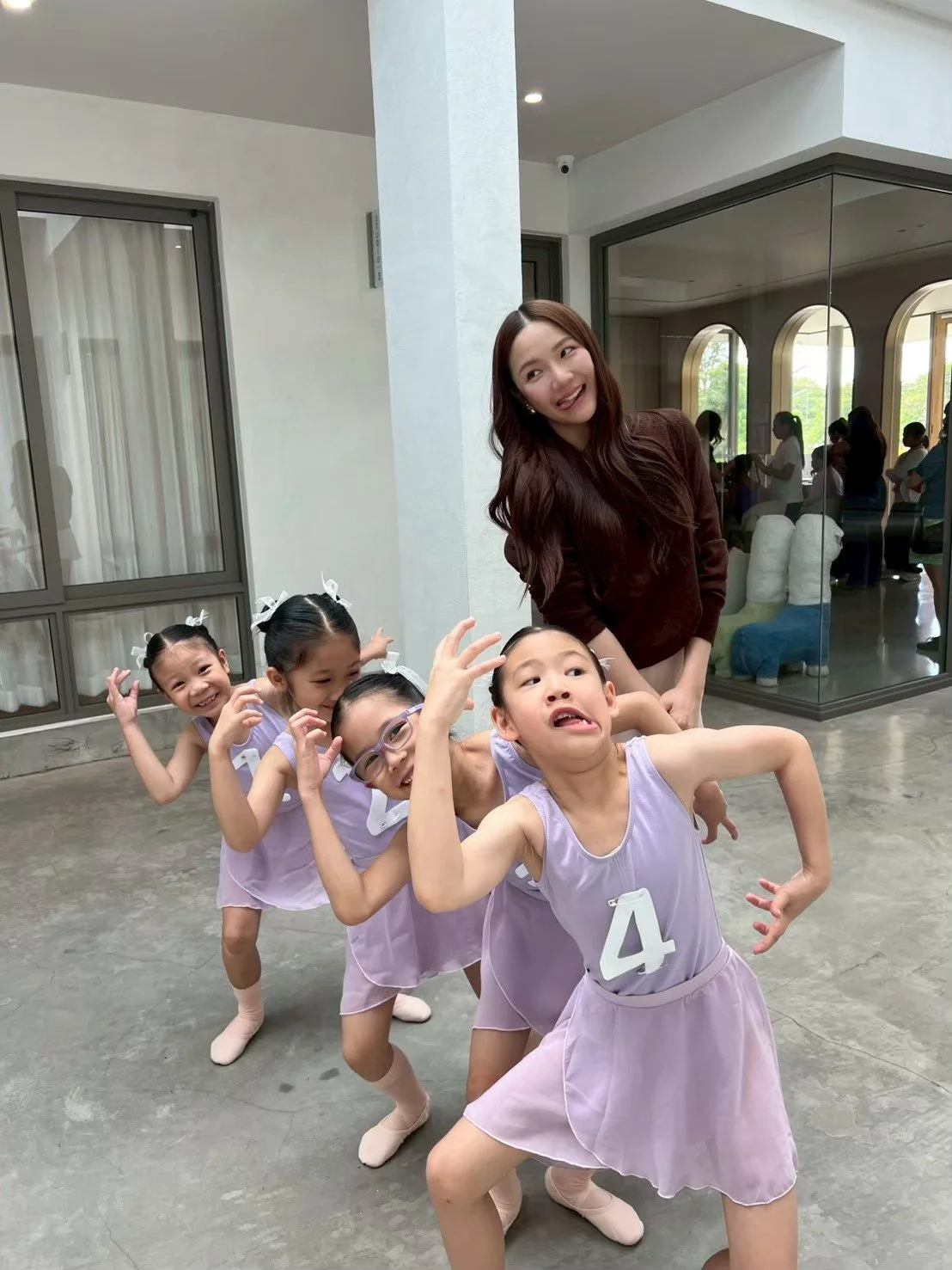 A woman with long brown hair and a brown top poses playfully with five young girls in lavender dance outfits, inside a modern building with large windows.