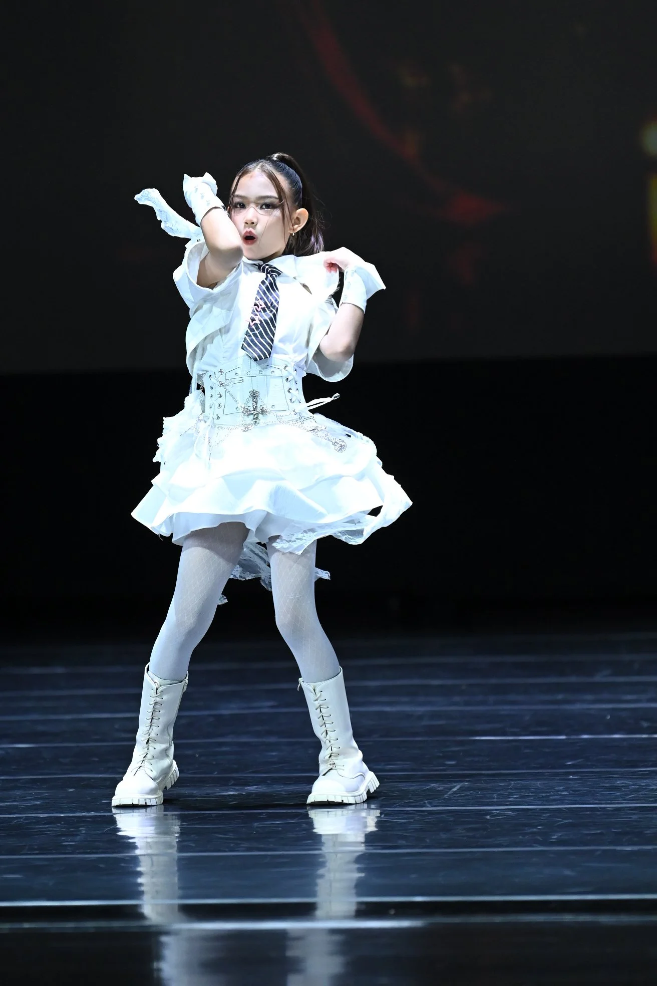 A young girl performing a dance on a dark stage, wearing a white dress with a plaid tie, white tights, and white lace-up boots, with glasses and a high ponytail.