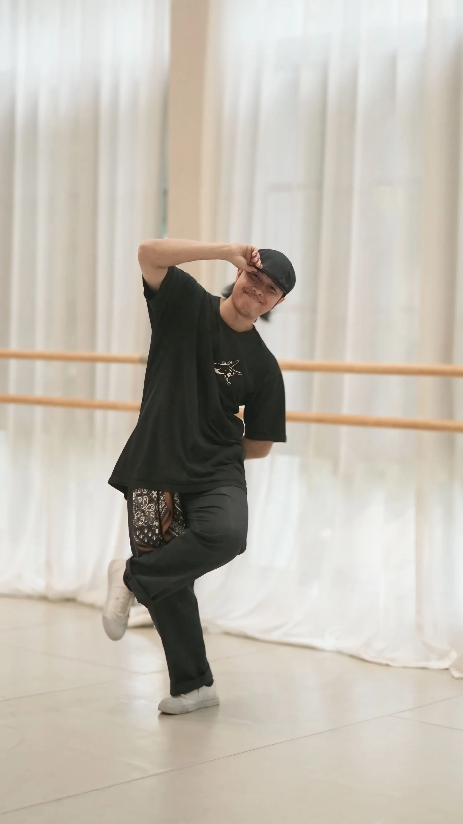 A young man in a dance studio wearing a black cap, black t-shirt, patterned shorts, and white sneakers, striking a dance pose with one knee bent and one hand near his head.