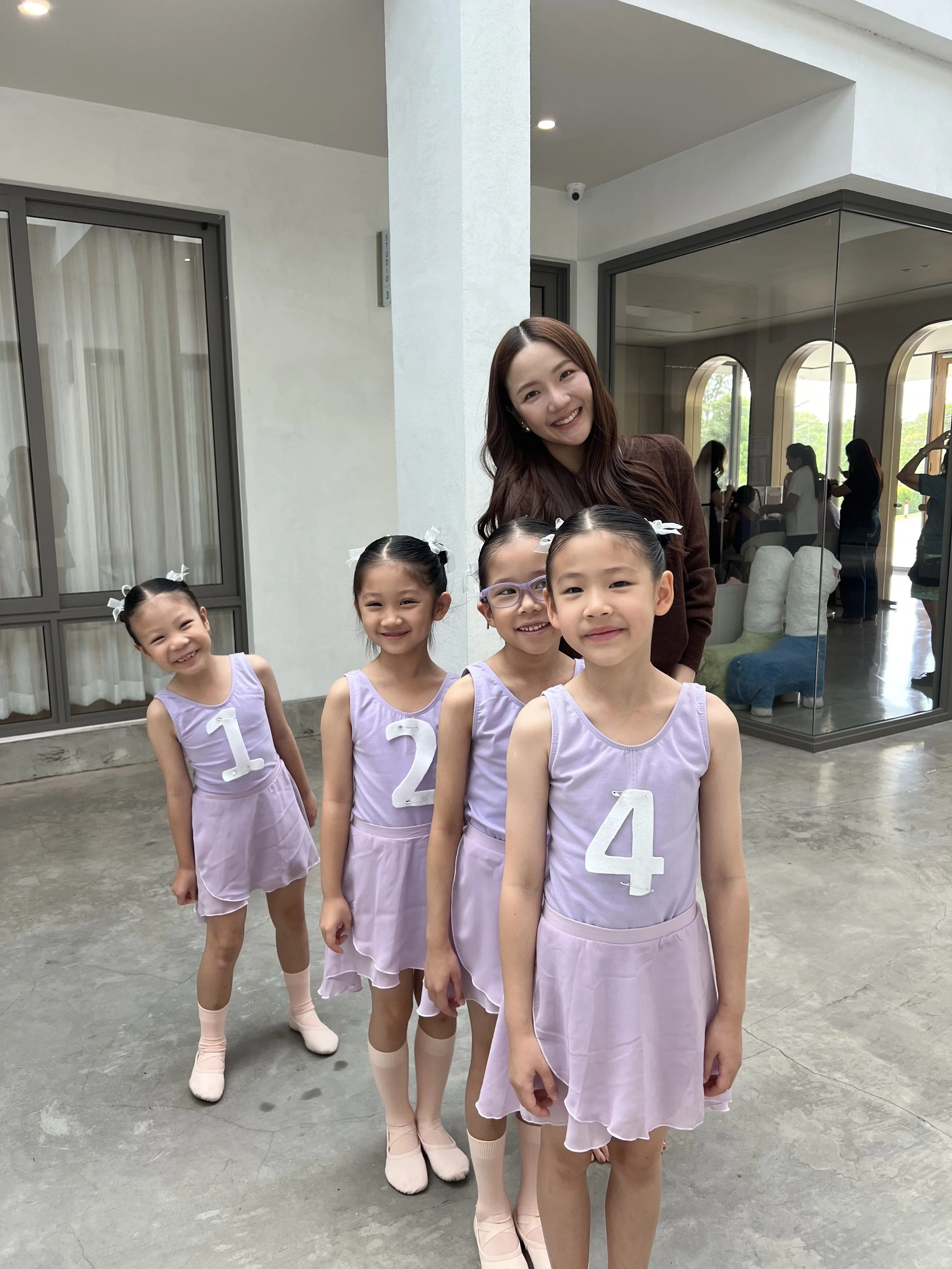 A group of four young girls dressed in light purple ballet outfits with white numbers on front, standing in a line with a smiling woman behind them. The girls have their hair tied back with white ribbons and are wearing ballet slippers. The backgroun