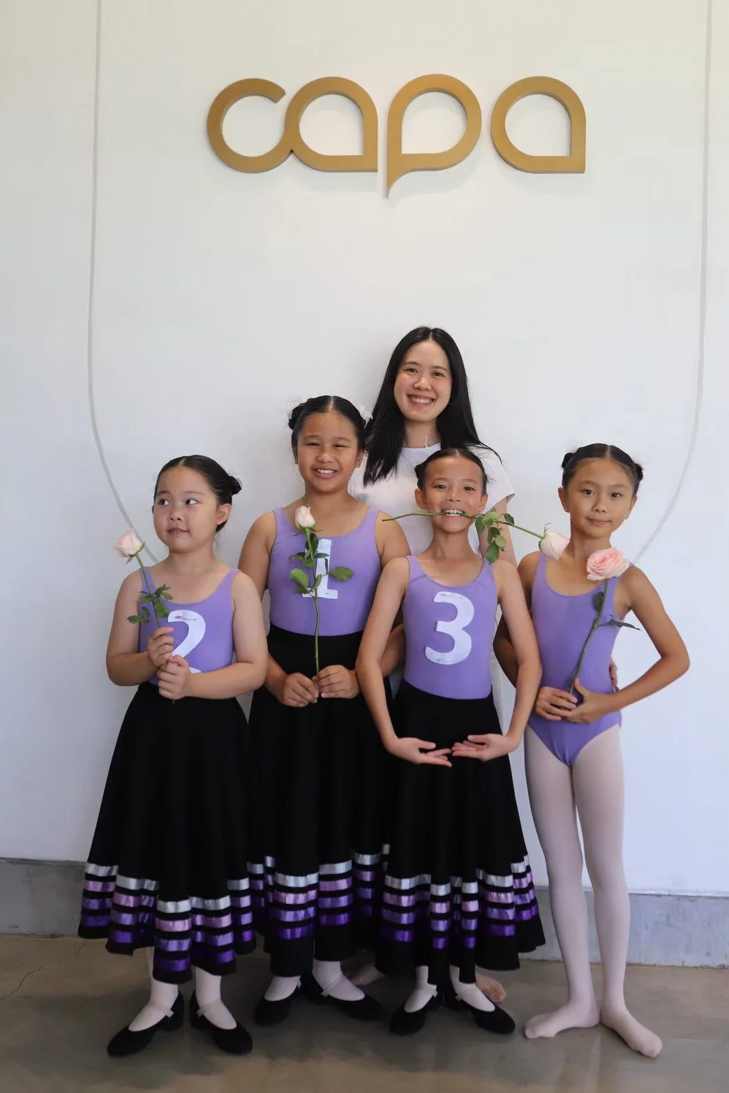 A group of five young girls and one woman standing in front of a white wall with a gold sign that says "capa." The girls are wearing purple leotards with numbers 2, 1, 3, and 4, and black skirts with purple stripes, holding pink and white flowers. Th