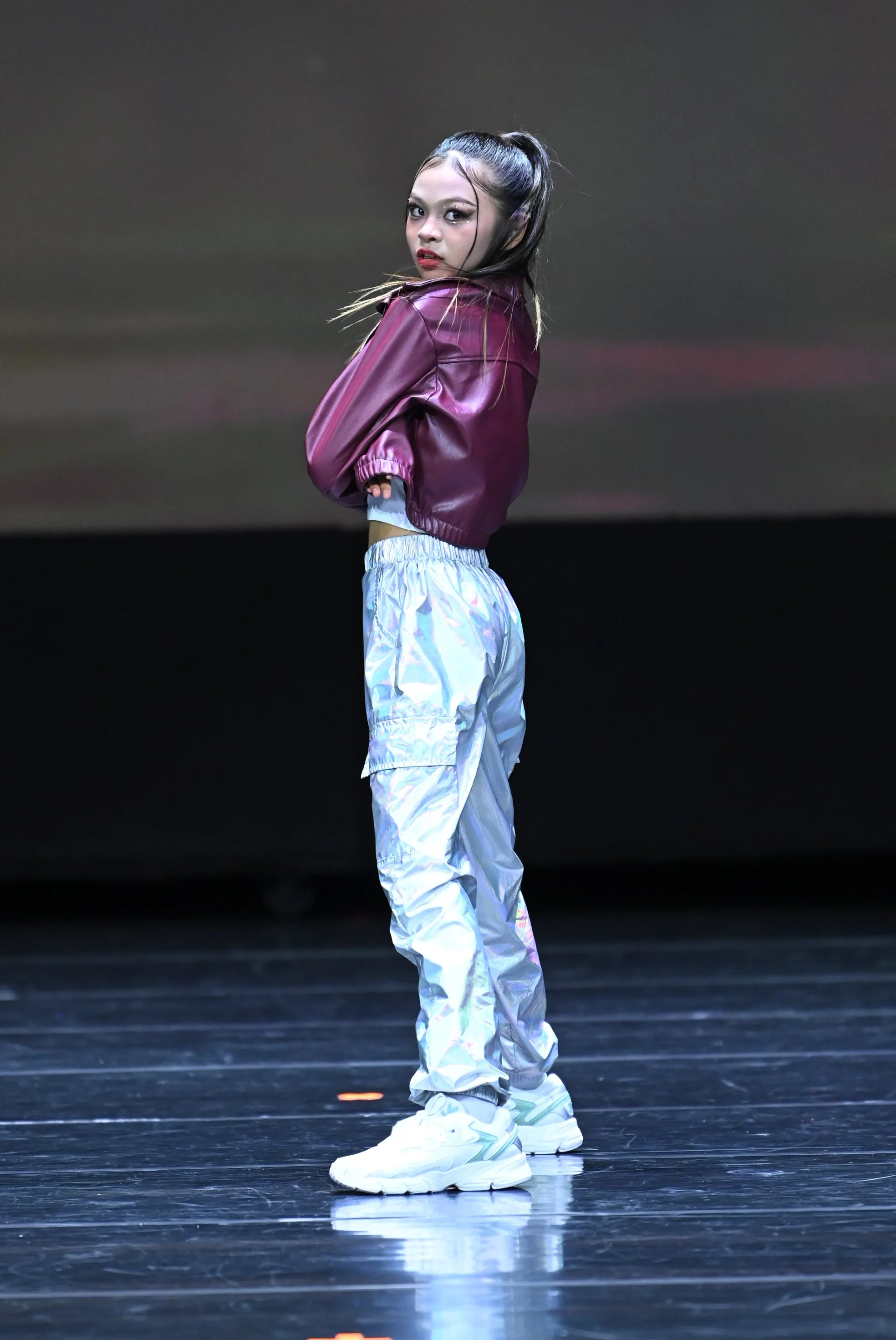 A young woman with long dark hair in a high ponytail, wearing a shiny purple jacket, white iridescent pants, and white sneakers, standing on a stage with a dark background, looking over her shoulder.