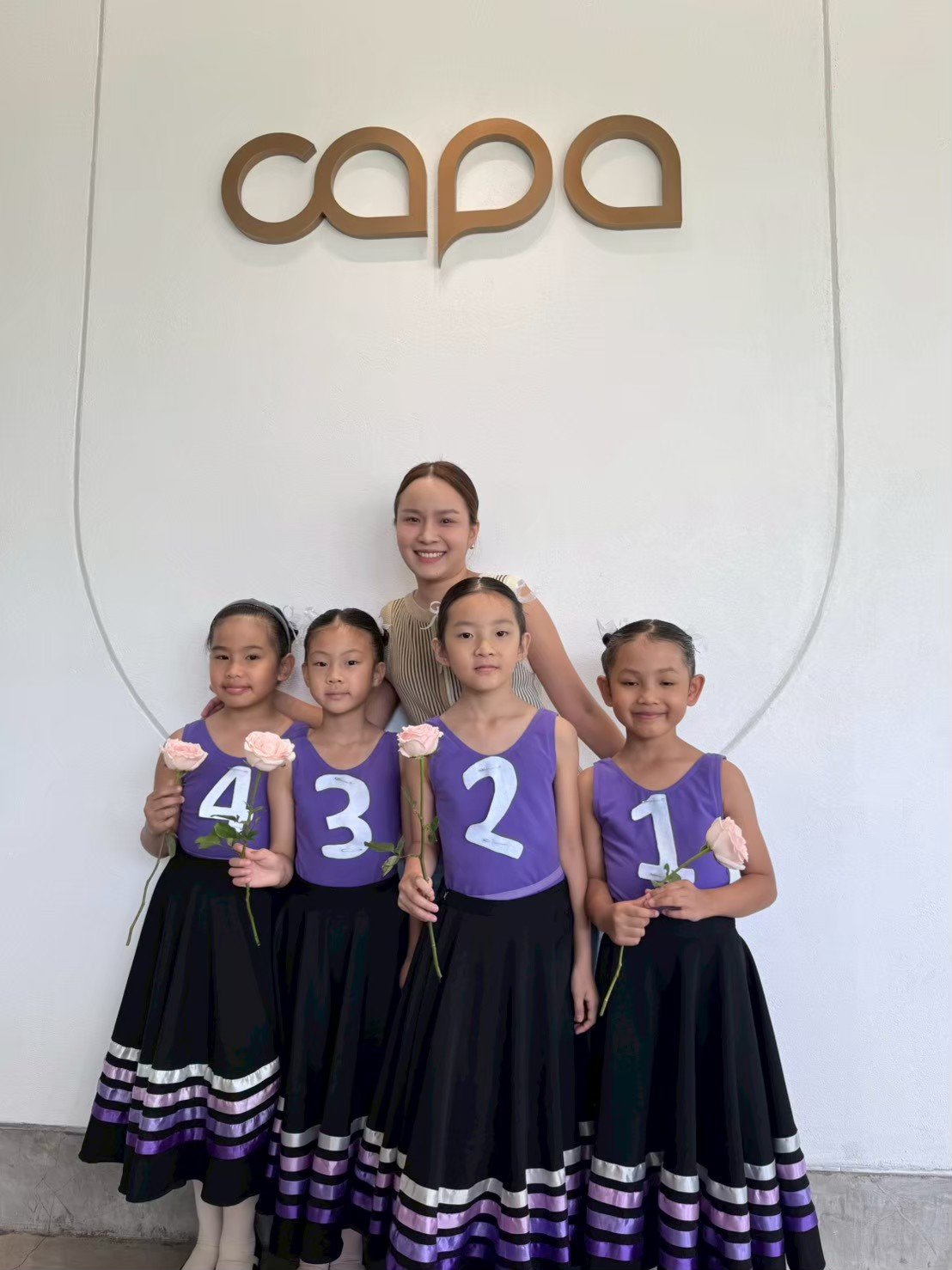 A group of five young girls and a woman standing together indoors in front of a white wall with the word 'capa' in large letters. The girls are wearing purple dance costumes with white numbers on their chests, holding pink roses, and the woman is sta