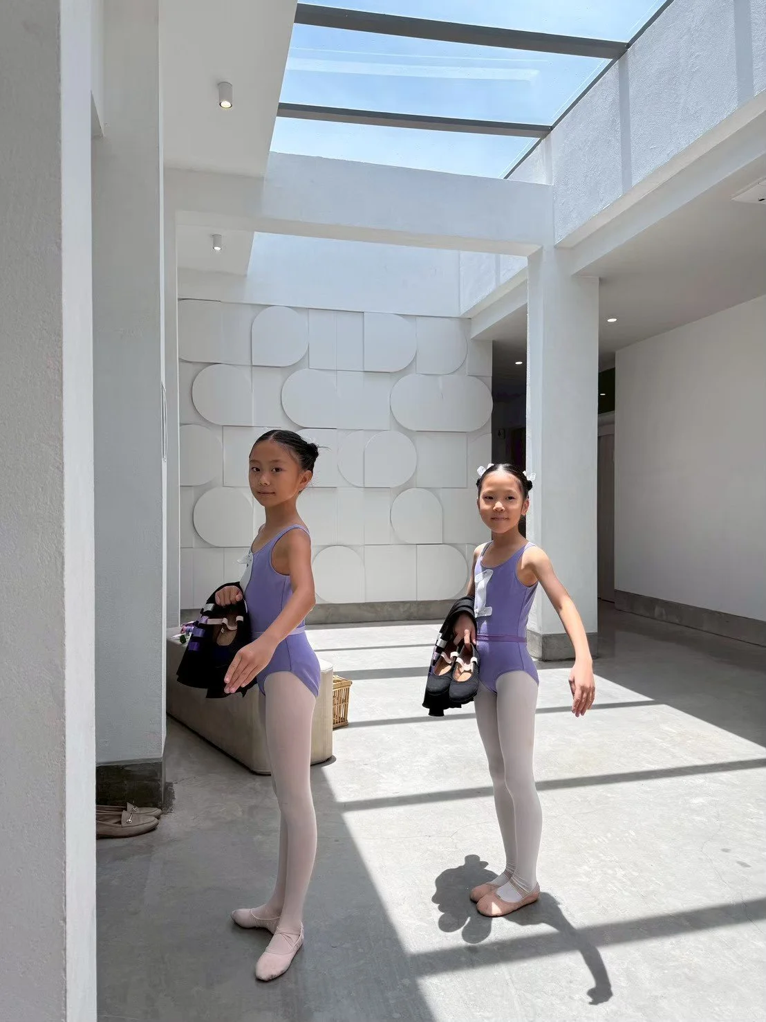 Two young girls in ballet leotards and tights standing in a modern, bright, white interior with high ceilings and skylights, holding ballet shoes and clothes.