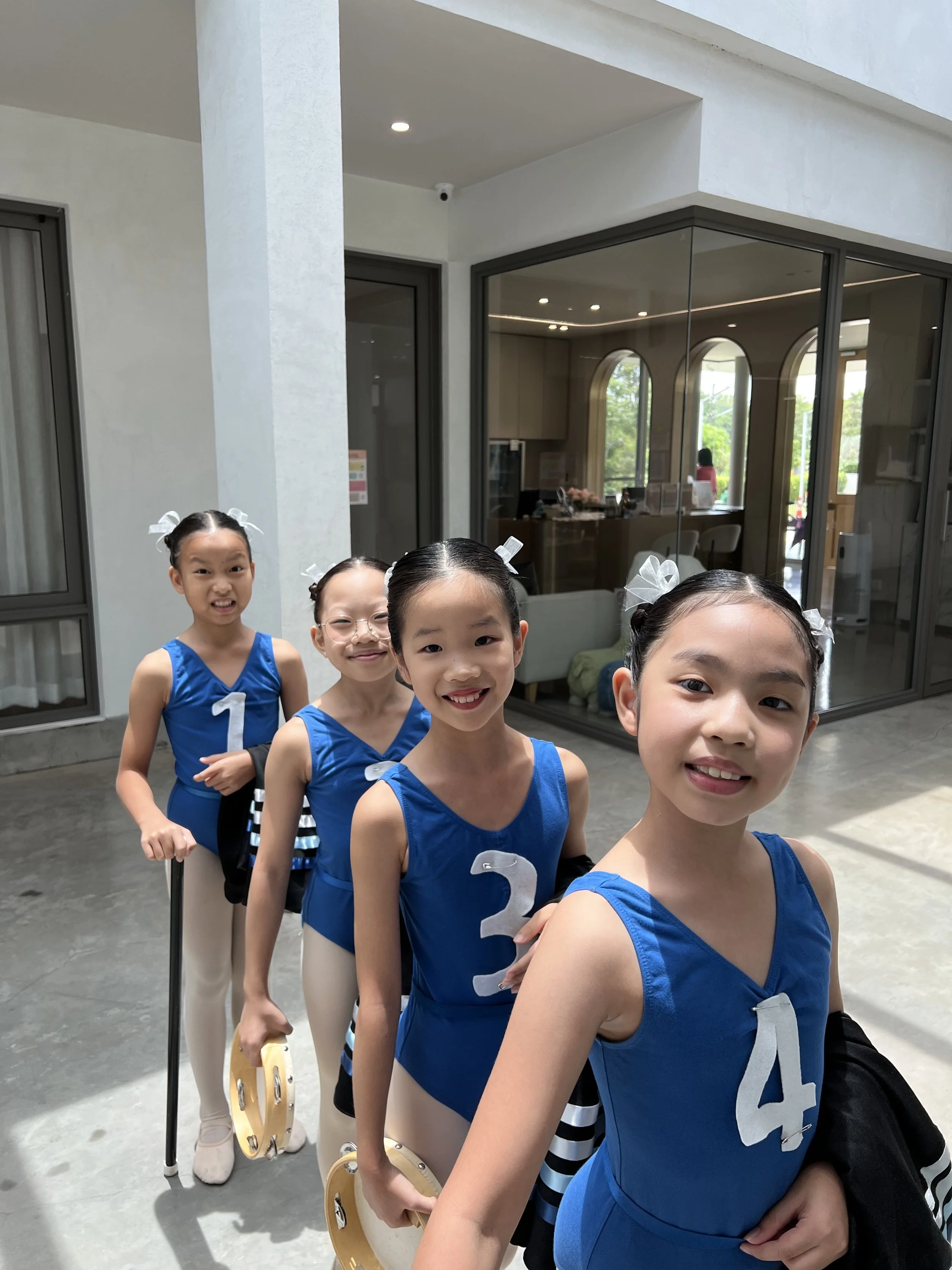 A group of young girls in blue cheerleading uniforms with white numbers, holding tambourines, standing in line inside a modern building with large windows and a concrete floor.