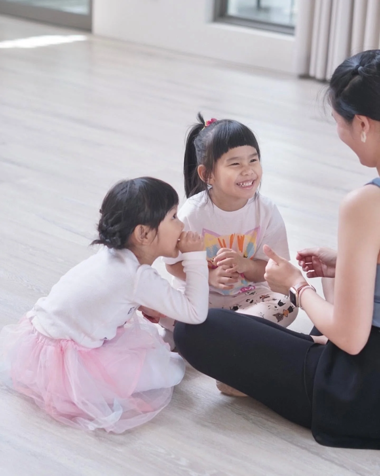 Two young girls sitting on the floor, engaging with an adult woman, smiling and laughing during a play or learning activity in a bright room.