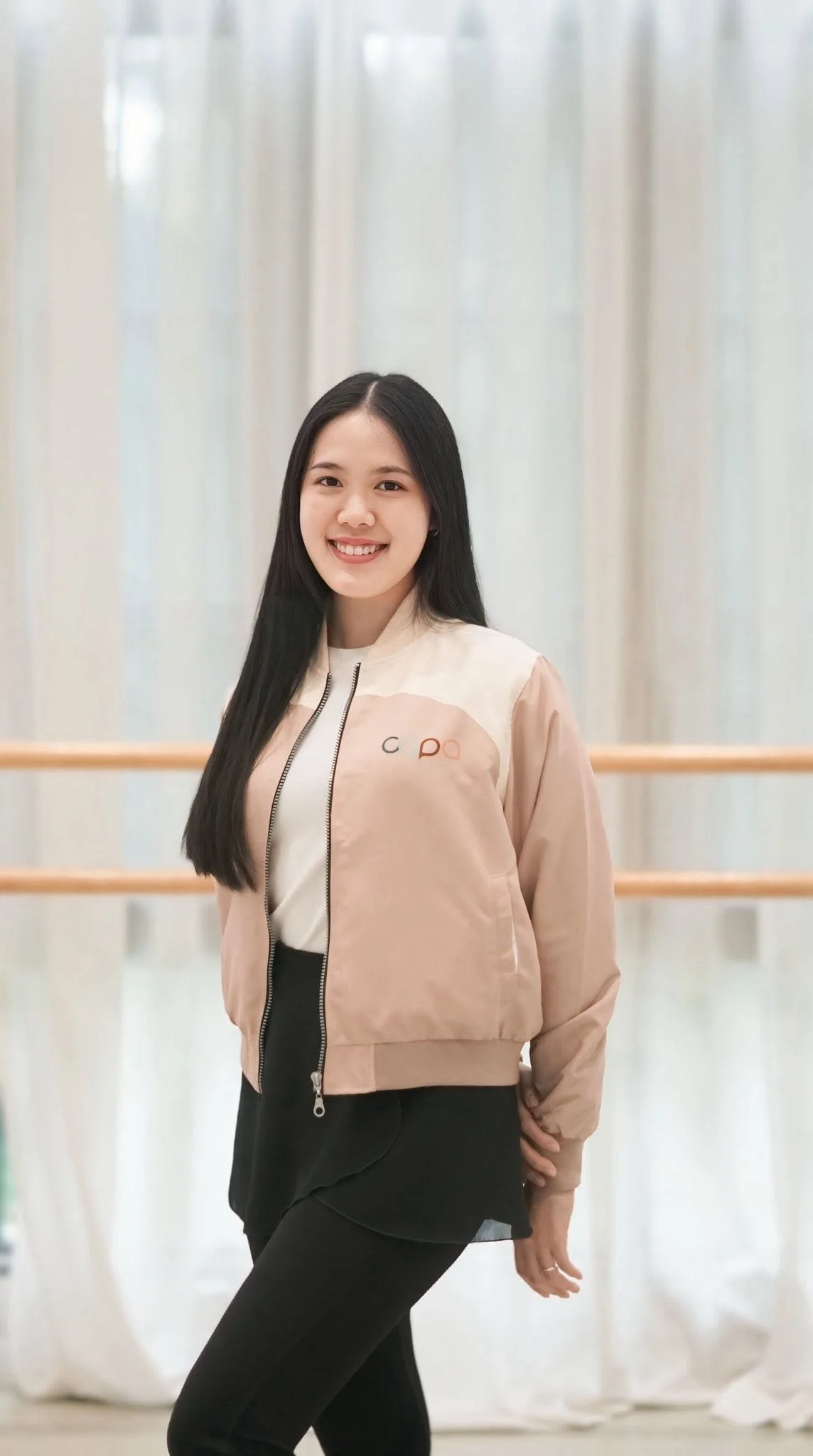 A smiling young woman with long black hair standing in front of light-colored curtains, wearing a beige jacket and black pants.