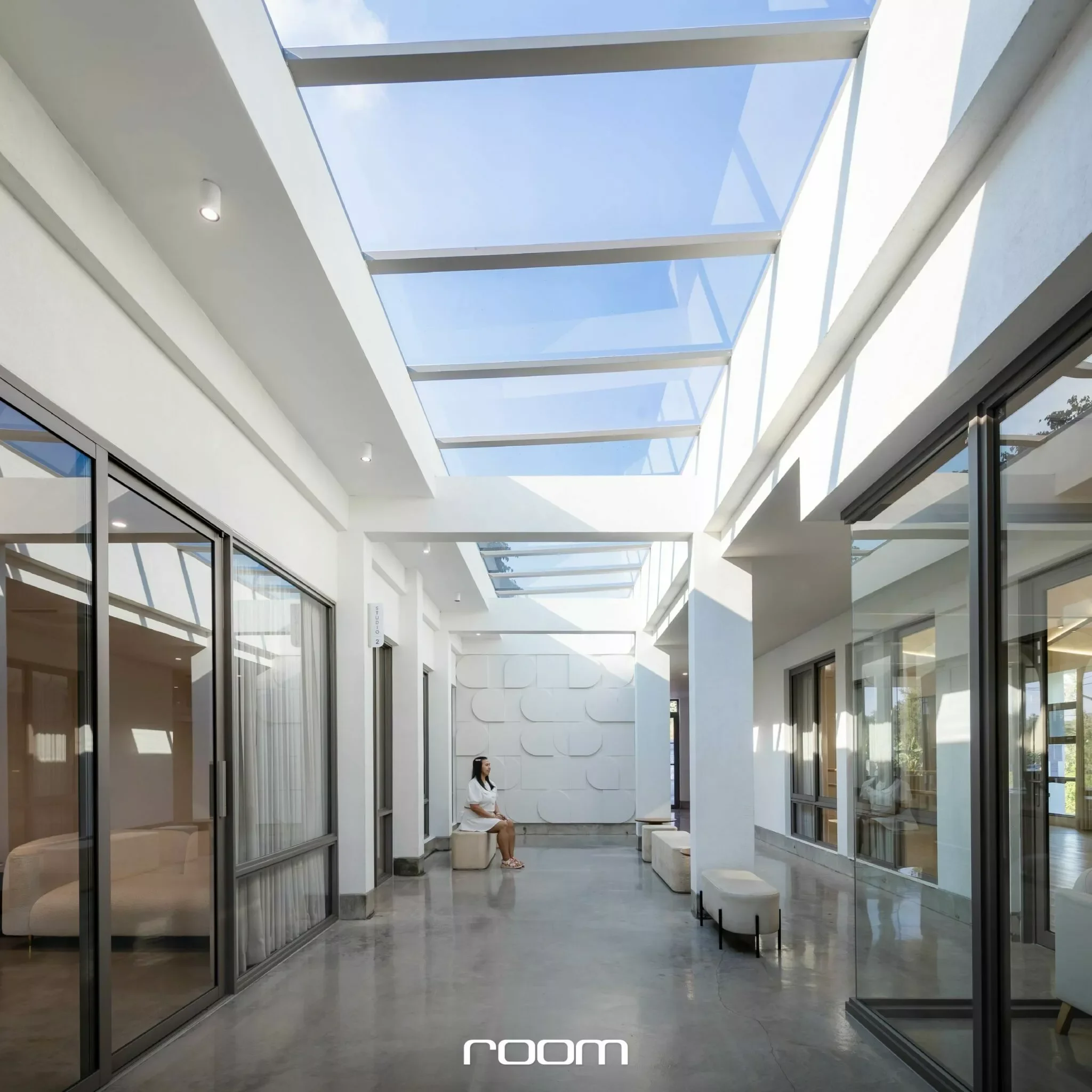 A modern indoor space with a high glass ceiling allowing natural light, white walls, glass doors, and benches. A woman is seated on one of the benches, and the floor is polished concrete.