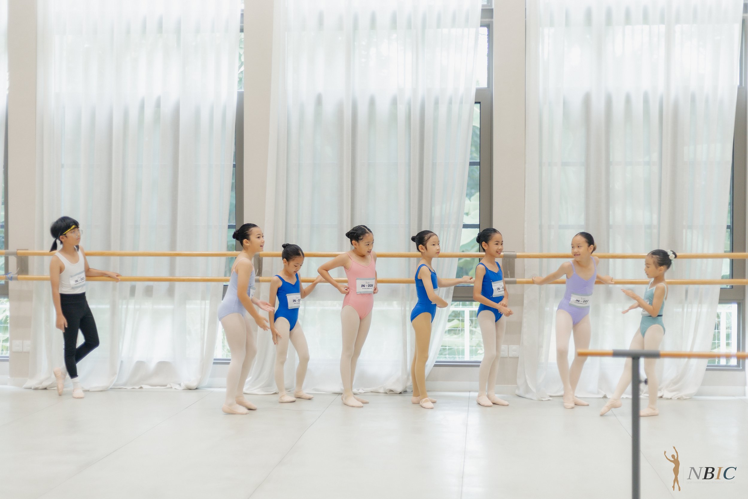 Young ballet dancers in practice at a dance studio with large windows and white curtains, wearing leotards and ballet slippers, some holding hands and talking.