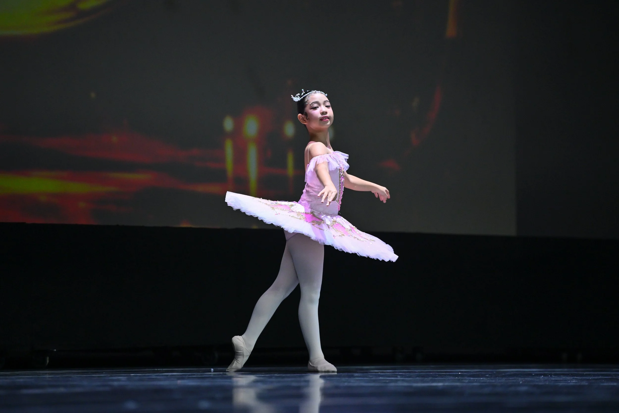 A young girl in a pink ballet costume with a tutu and a tiara performing ballet on stage.