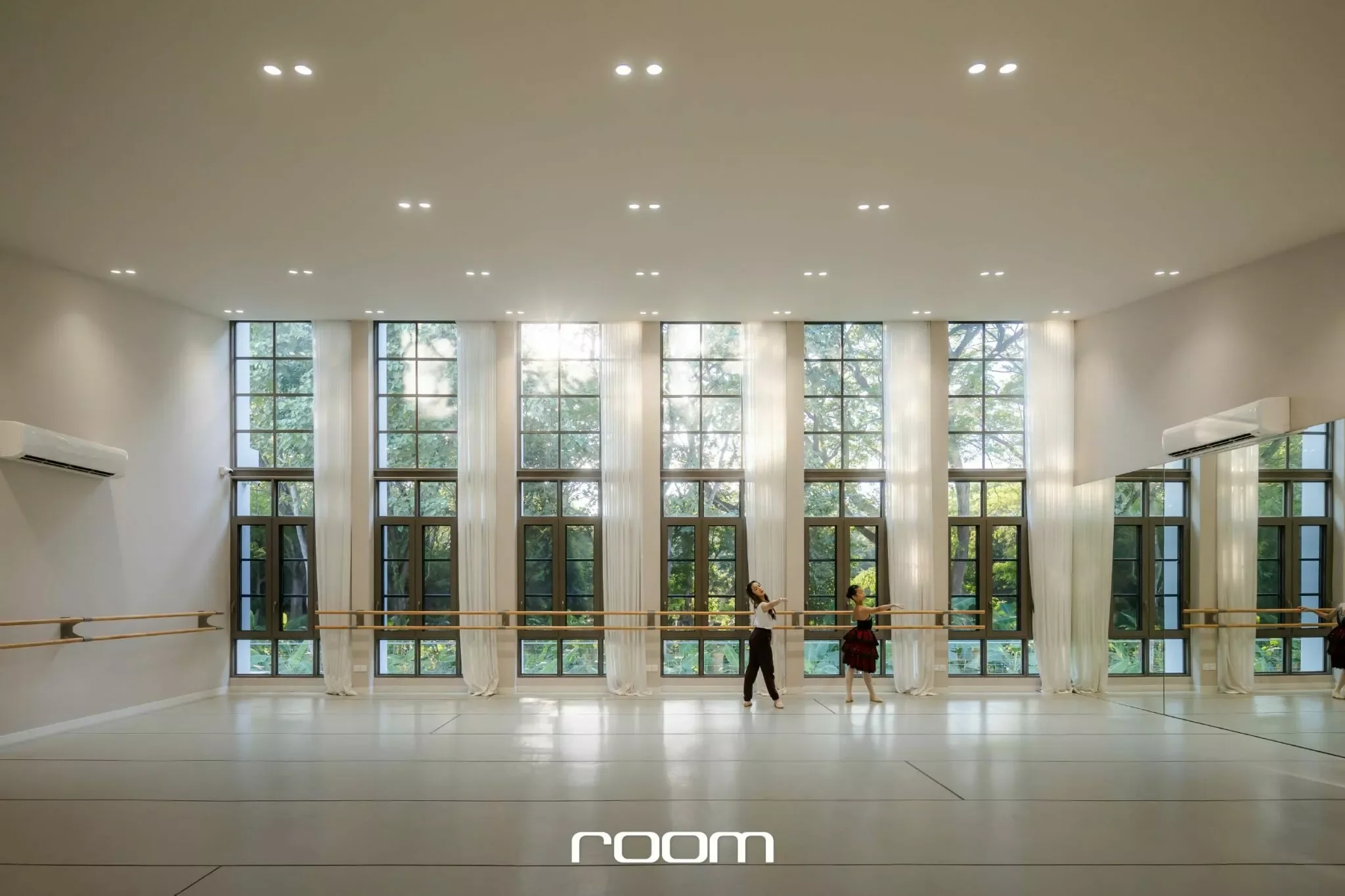 A spacious dance studio with large windows, white walls, and ceiling lights. Two women are practicing ballet at the barre.