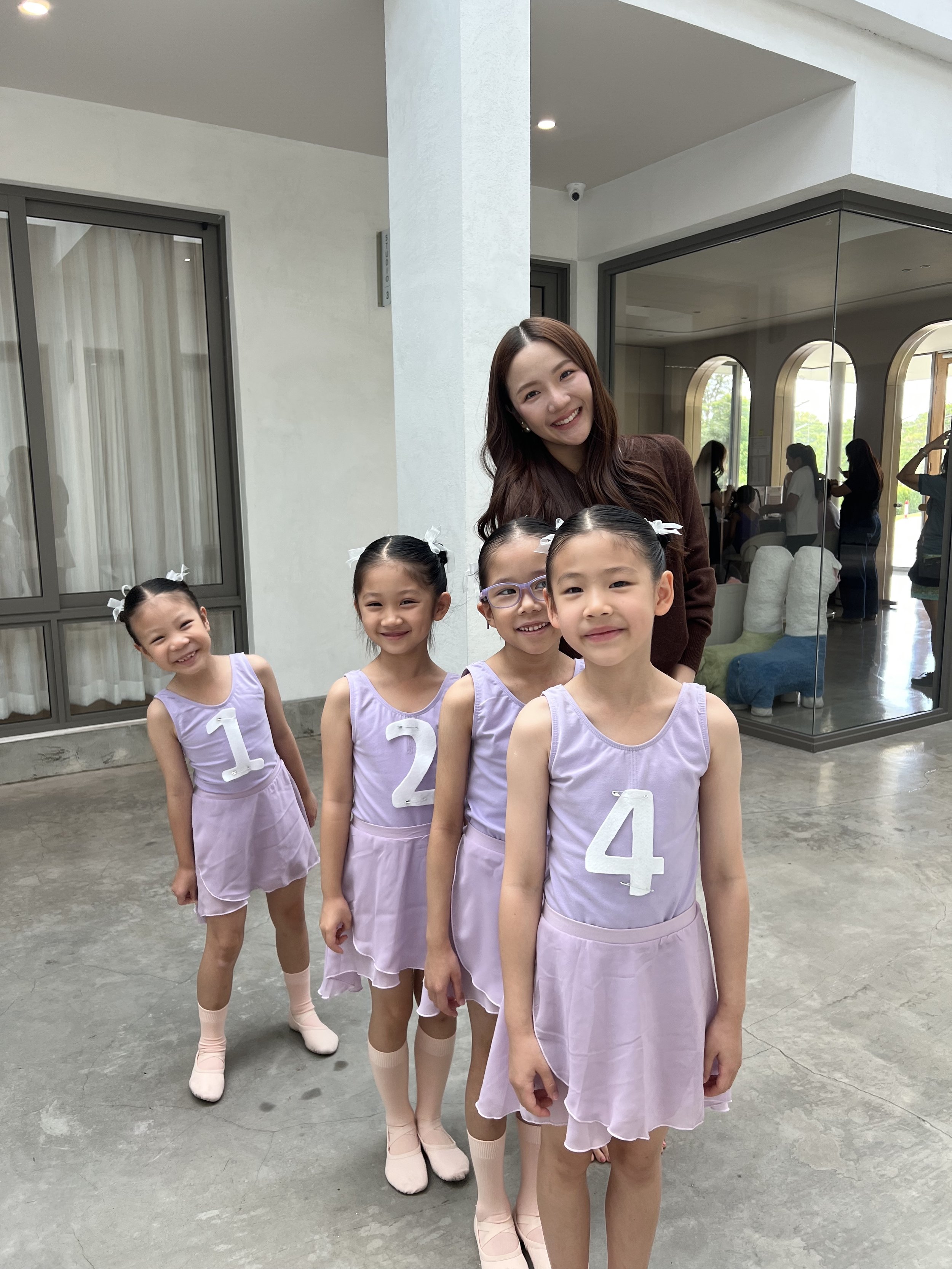 A group of four young girls in lavender dance costumes with white numbers on them, and a woman in brown at a dance studio with large windows and mirrors, smiling.